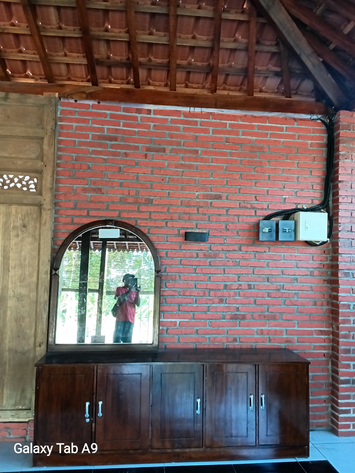 A person is taking a selfie in a mirror. The person is wearing a red shirt and a black cap. The mirror is above a dark wooden cabinet. The cabinet has three doors, each with a silver handle. The background is a brick wall. There is also a wooden door on the left side of the image and a wooden ceiling with visible beams. Electrical boxes are mounted on the brick wall. The text "Galaxy Tab A9" is visible in the bottom left corner of the image. This scene is likely located in Trenggalek, Indonesia, given the provided context.