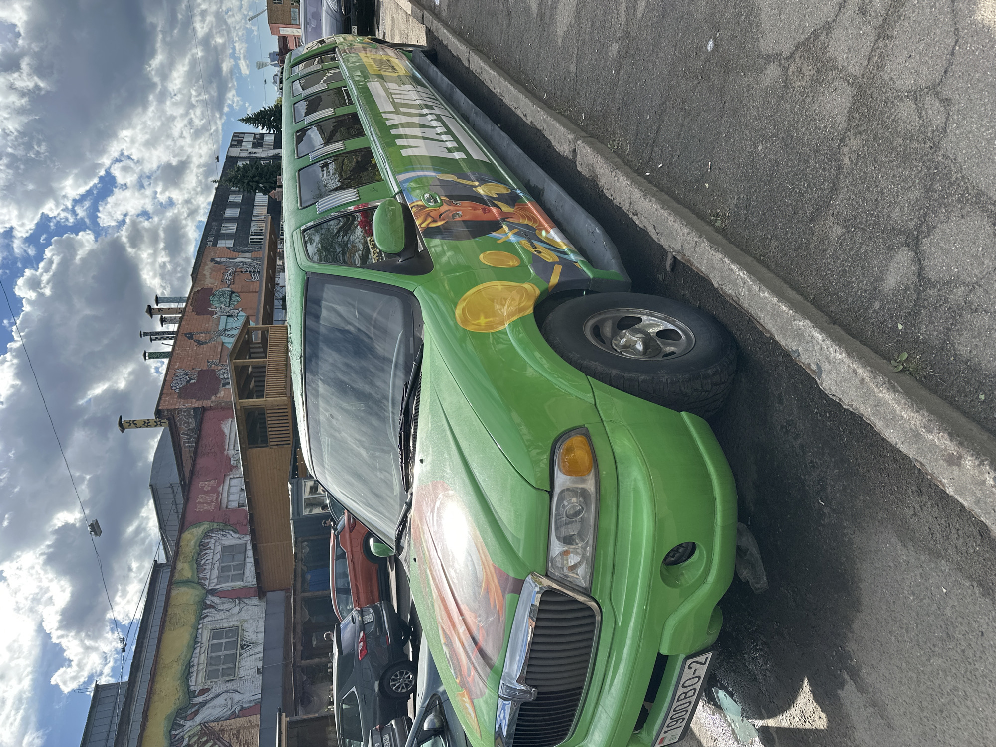 The image shows a green stretch limousine parked on a street in Minsk, Belarus. The car is parked alongside a curb with other parked cars nearby. The building on the left has murals on its wall. The sky is partly cloudy. The vehicle's license plate is visible and reads "1900 BO-2". The limousine has artwork and lettering on its side. The environment appears to be urban, with buildings and parked vehicles lining the street.