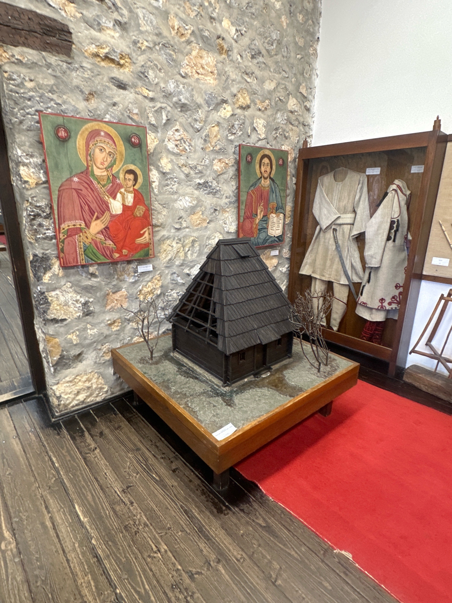 The image displays an indoor exhibit within the city of Topola Municipality, Serbia. The exhibit features a scale model of a traditional Balkan house with a dark, multi-tiered roof, placed on a base resembling a landscape. Two religious icons hang on a stone wall behind the model; one depicts the Virgin Mary and Christ Child, and the other shows Jesus Christ. To the right, a glass-fronted display case contains traditional Serbian clothing, including tunics and embroidered garments, along with a sword. The floor is made of dark, wooden planks, and a vibrant red carpet runs along the right side of the exhibit. The overall scene suggests a cultural or historical museum setting.