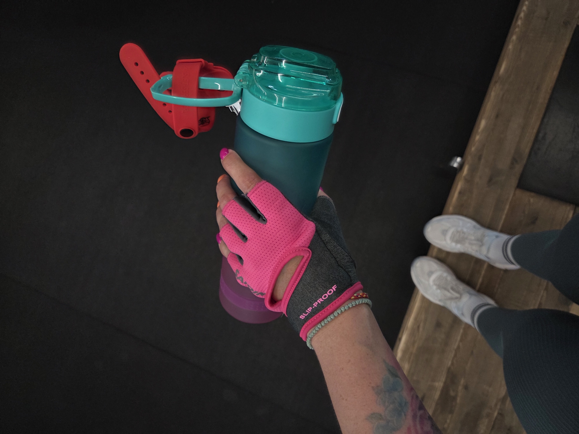 The image shows a person holding a water bottle while standing on what appears to be a wooden platform next to a black floor. The water bottle is dark teal with a lighter teal lid and a red attachment mechanism. The person is wearing a workout glove with pink accents and a grey back, labeled "SLIP-PROOF." The person has colorful nail polish and bracelets. Only the person's arm, legs, and shoes are visible. The legs are covered in dark pants and white sneakers.
The location in the city of Birobidzhan, Russia, cannot be determined from the provided image.