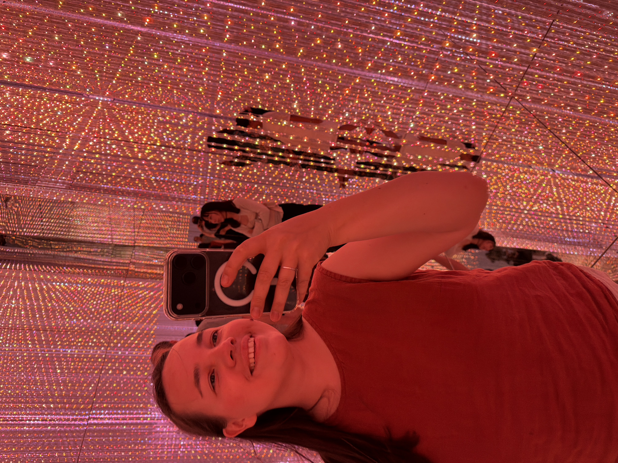 A young woman with brown hair is taking a selfie in a room filled with red and orange lights. She is wearing a red sleeveless top and a ring on her left hand. In her right hand, she holds a smartphone, also capturing the reflection of other people and the light installation in the background. The environment appears to be an art installation, possibly the "teamLab Borderless" digital art museum in Koto, Japan, known for its immersive light displays. The background is a dense arrangement of small lights, creating a shimmering and colorful effect.