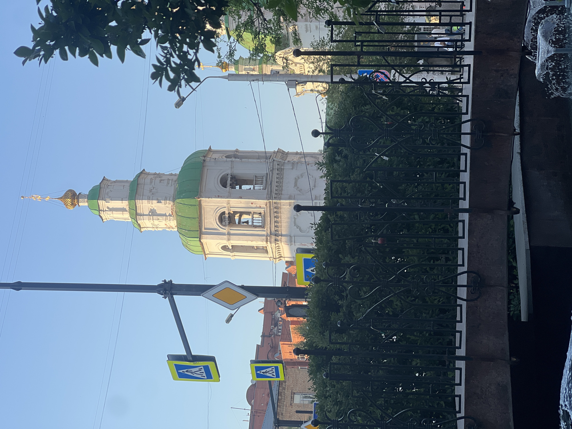 The image shows a view of the Annunciation Cathedral in Krasnoyarsk, Russia, partially obscured by trees, a fence, and street signs. The cathedral's tiered tower with its green domes and golden cross is the focal point. A lamppost and pedestrian crossing signs are visible in the foreground. The sky is blue. The fence is black metal. A building of a reddish-brown color is further in the distance.