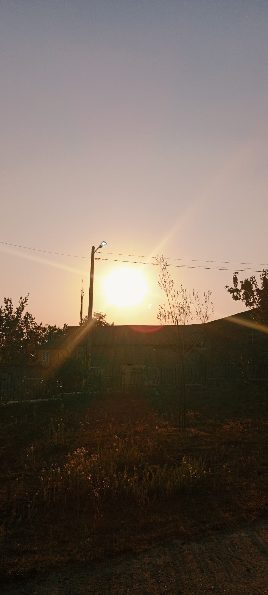 The image depicts a sunset scene in the city of Adai, Spain. A bright sun is visible in the sky, casting warm light and lens flares. In the foreground, there is dry grass and vegetation, with a dark, possibly gravel, surface in the lower right corner. Behind the vegetation, a fence runs horizontally across the frame. A building with a dark roof and a lighter colored wall is visible behind the fence, with a window on the left side. A street light on a pole is positioned to the left of the sun, with power lines stretching across the sky. To the right of the street light and partially obscuring the sun, a thin tree with sparse leaves is visible. The sky is a gradient of soft pink and pale blue, indicative of twilight. The overall impression is one of a rural or suburban landscape at dusk.