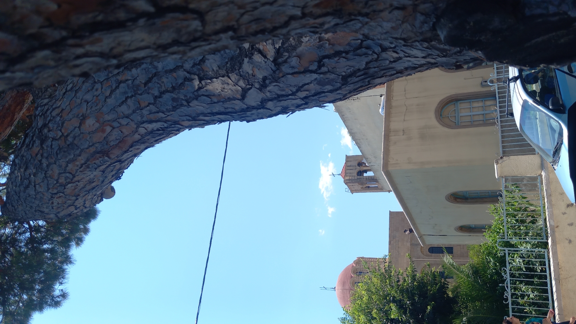 The image shows a view from behind a large tree trunk looking towards a bright blue sky with a few scattered clouds. In the middle ground, there are several buildings, including a church with a red dome topped with a cross, and a bell tower with a cross. The buildings are made of light-colored stone and have arched windows. A metal fence runs in front of the buildings. A light blue car is parked in the foreground. The scene appears to be located in a sunny, urban environment. Based on the architectural style and the presence of a church, this is likely a street view in the city of Kos, Greece.