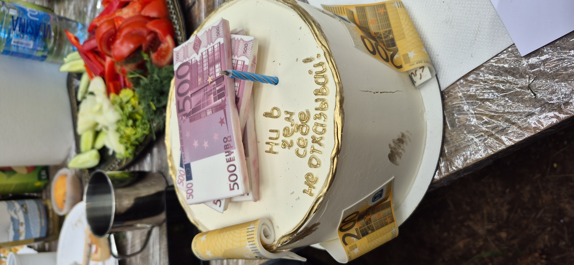 The image captures a celebratory scene, likely a birthday, set outdoors with a focus on a custom-decorated cake. The cake is round, covered in white frosting with a gold-trimmed edge, and adorned with edible decorations resembling euro banknotes of various denominations (500 and 200). A partially burnt blue and white striped candle sticks out among the "500 euro" bills. Inscribed on the cake's side in gold lettering is a phrase written in Russian which translates to "Why deny yourself anything!".
Beside the cake is a platter with chopped vegetables, including red peppers, cucumbers, and greenery (possibly dill). To the left of the platter, there's a stack of metal camping-style cups and a bottle of mineral water.
The environment suggests an outdoor setting, possibly a picnic, with the cake placed on a surface covered with wrinkled plastic film over a dark wooden table. The location appears to be Mali Bostur, Montenegro, based on the text included.