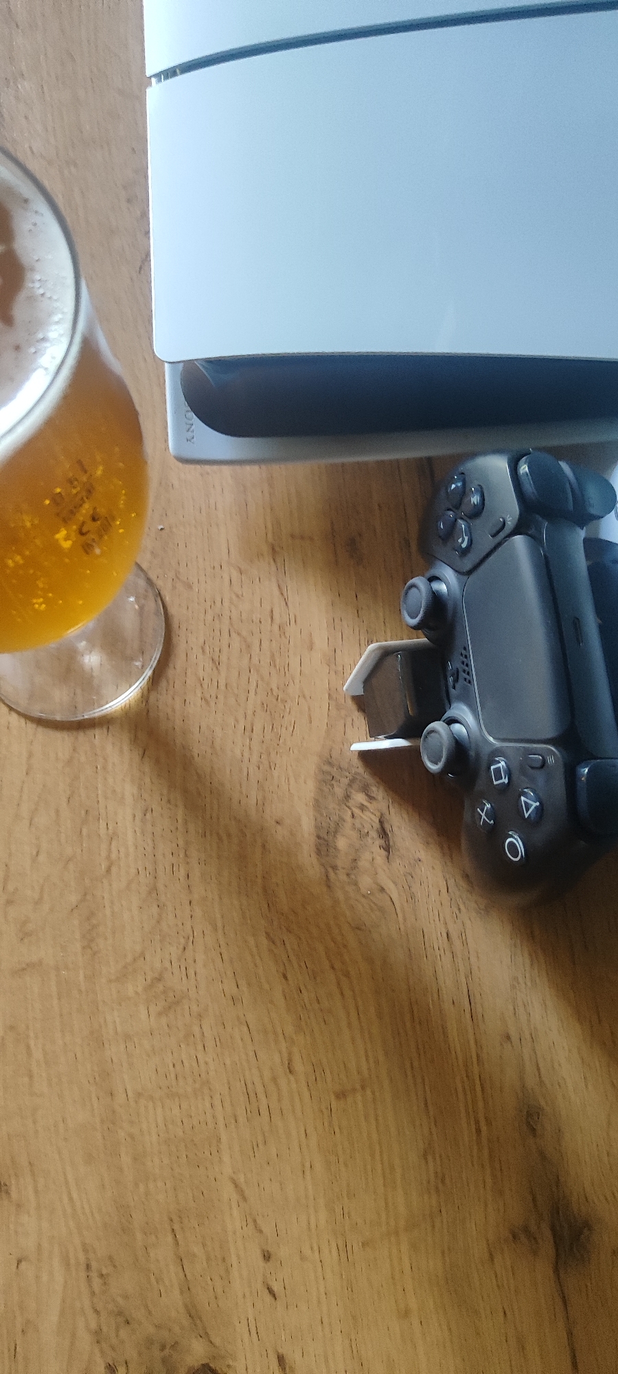 A glass of beer and a video game controller rest on a wooden table next to a white game console. The scene is indoors, and the lighting suggests it is daytime. There are no people visible in the image.