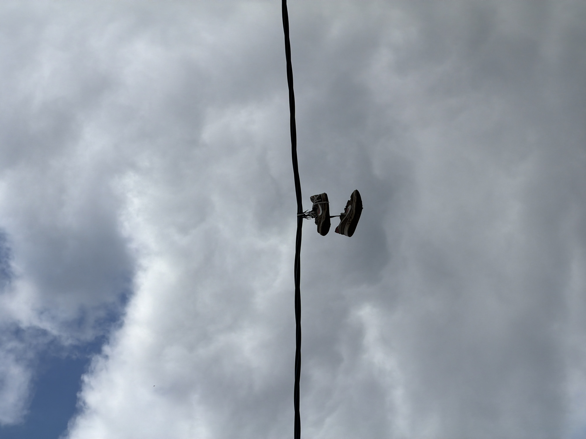 A pair of worn athletic sneakers, featuring tan or light brown uppers and white or light-colored soles, are tied together by their laces and suspended over a thick, dark, horizontal utility cable. The shoes hang centrally beneath the cable, with one shoe positioned directly above the other. The background is a predominantly overcast sky composed of dense gray and white stratocumulus clouds, revealing a small patch of blue sky in the upper left quadrant. No individuals or additional structures are present. This scene is located within the city of Krasnodar, Russia.