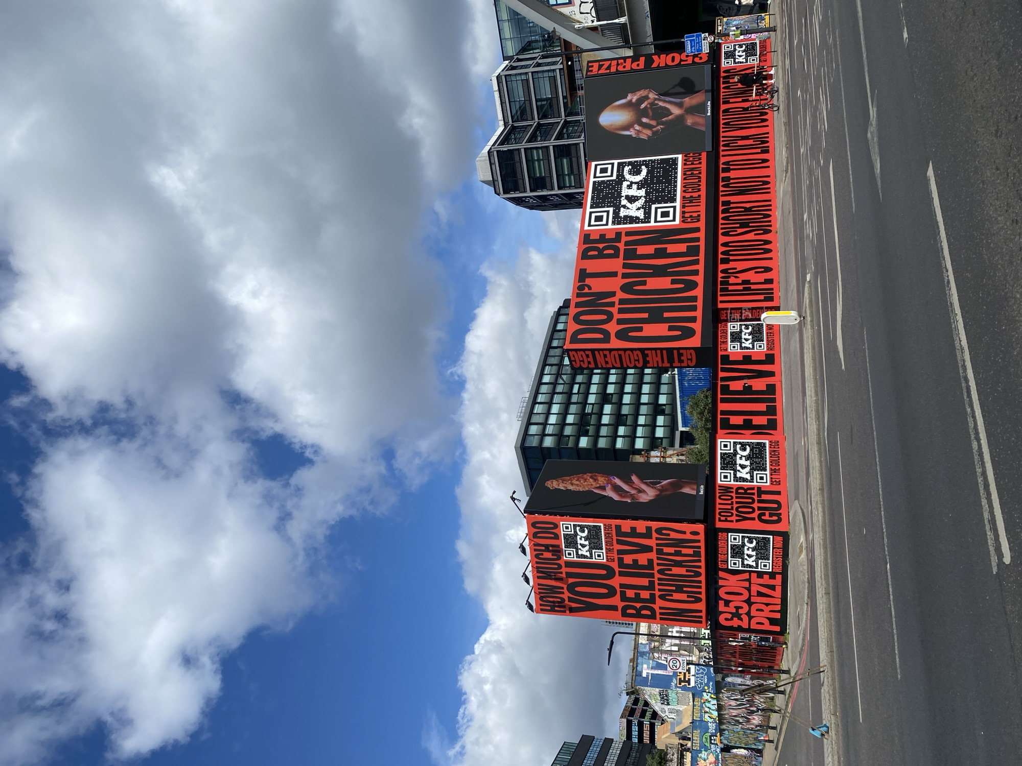 The image depicts a street scene in London, United Kingdom. A series of large KFC advertisements are positioned along the side of the road, displaying phrases such as "HOW MUCH DO YOU BELIEVE IN CHICKEN?", "DON'T BE CHICKEN", and "LIFE'S TOO SHORT NOT TO LICK YOUR FINGERS", along with QR codes and images of KFC products. The ads are mounted on what appears to be a temporary structure or fence. The road is asphalt, and white lane markings are visible. Buildings with modern architecture and glass facades form the background, along with a blue sky partially covered with white clouds. Graffiti-covered walls can be seen in the lower part of the frame. There is no visible human presence in the shot.