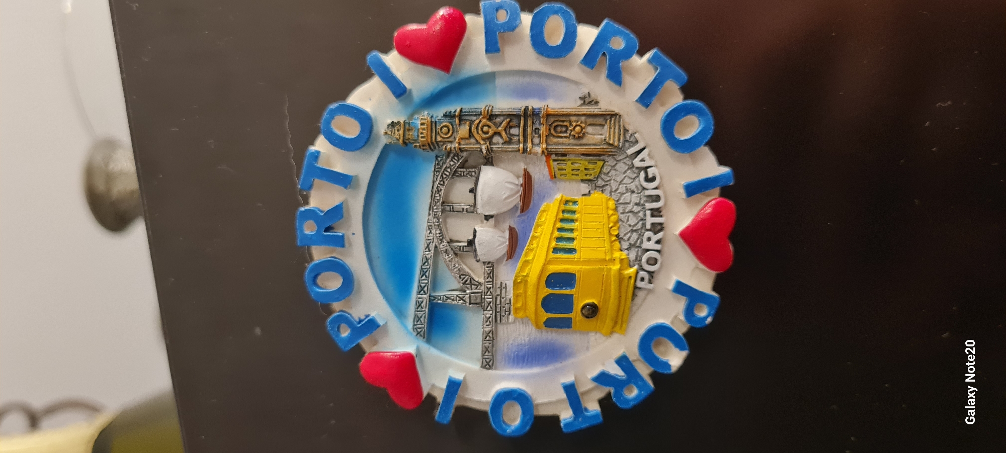 This is a close-up, angled shot of a circular refrigerator magnet. The magnet is predominantly white with blue lettering and red heart accents. The lettering spells out "I <3 PORTO PORTUGAL" in a circular pattern. In the center of the magnet, there is a raised, colorful depiction of a cable car or funicular, with a yellow car and a gray structure with white domes. The background of the magnet is a gradient of blue and white, possibly representing water and sky. The magnet is attached to a dark, glossy surface, likely a refrigerator door, as evidenced by the reflection of a light source on the surface and a small, out-of-focus object to the left. The bottom right corner of the image contains text that reads "Galaxy Note20," indicating the device used to capture the photo. There are no people visible in the image. The scene appears to be indoors, given the artificial lighting suggested by reflections. The magnet itself is the sole focus of the image, showcasing a souvenir from Porto, Portugal.