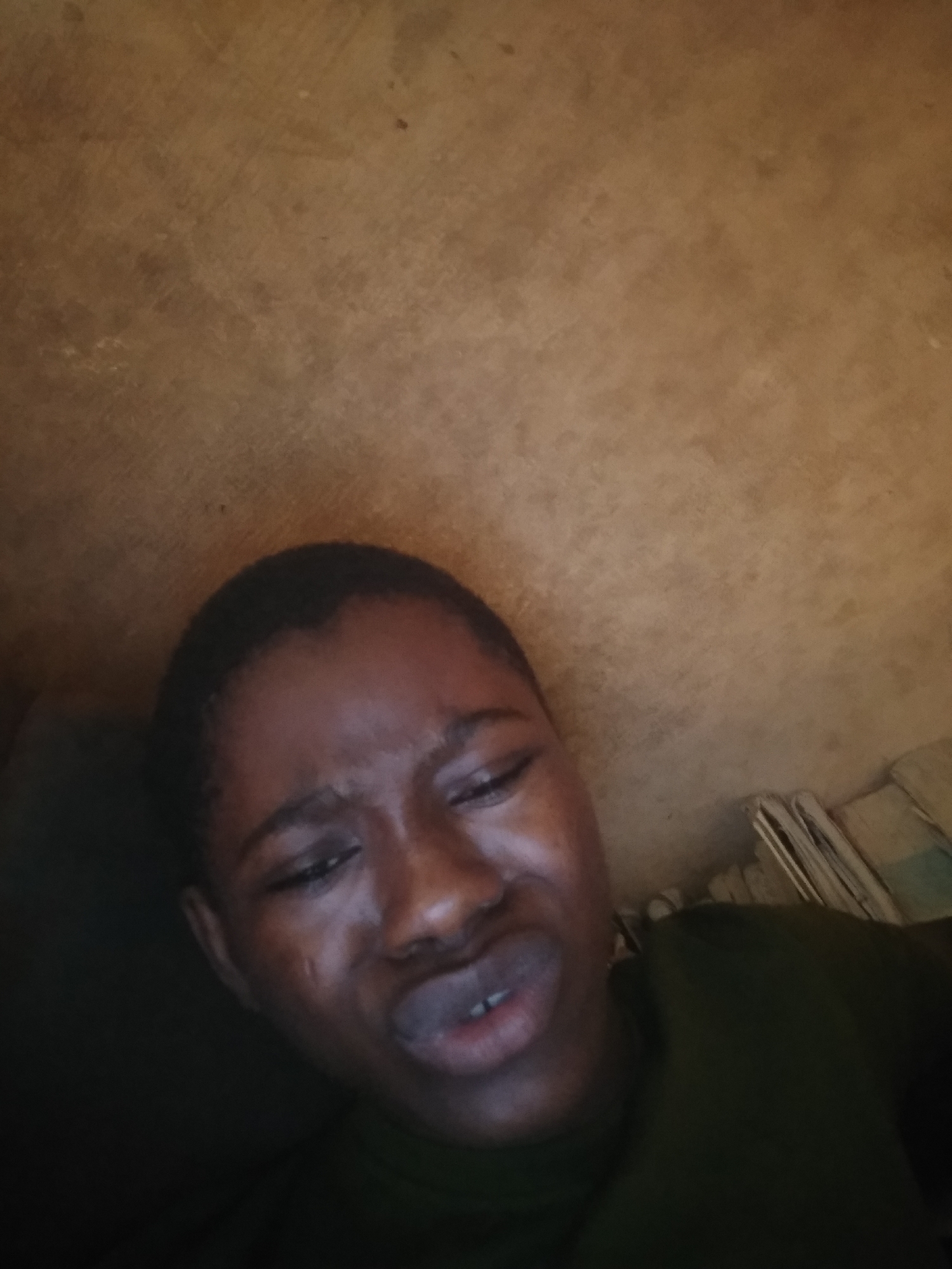 A close-up, eye-level shot depicts a person with dark skin, a shaved head, and facial contortions suggesting distress or discomfort. The background is a textured, light-brown surface, possibly a wall. To the lower left, a dark area is present, indicating a possible pillow or cushion. To the lower right, stacked items are visible, possibly papers or books. The person is wearing a dark-green shirt. The location is listed as Unknown, Unknown.