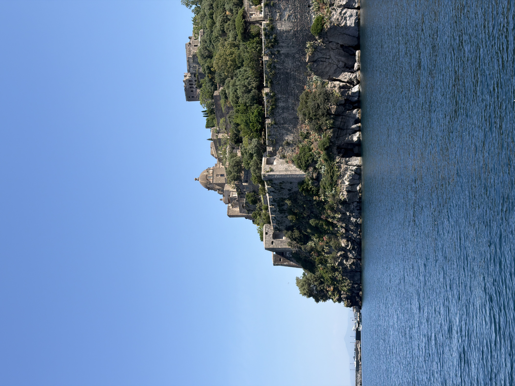 The image depicts the Aragonese Castle in Ischia, Italy, perched on a rocky islet connected to the main island by a causeway. The left of the frame shows a clear blue sky, while the right side reveals the blue waters surrounding the castle. The castle itself is built into the rock face, with medieval architecture including walls, towers, and a domed church visible amidst lush green vegetation. The image is oriented vertically, emphasizing the castle's height and its dramatic location on the water's edge.