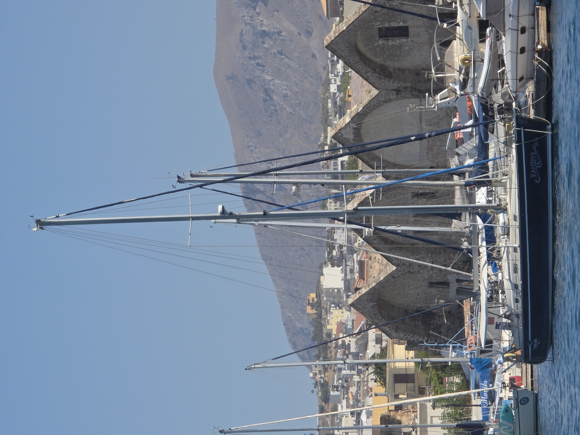 In Chania, Greece, a tranquil harbor scene unfolds under a clear blue sky. Dominating the foreground are several sailboats, their masts tall and slender, creating a striking silhouette against the backdrop of a Mediterranean town. The largest sailboat, a dark hull with "Kamisto" inscribed in white lettering, is moored at the front, showcasing its elegant design. In the mid-ground, traditional stone buildings with arched openings, reminiscent of Venetian architecture, stand as a testament to the city's rich history. Beyond these structures, a densely populated town sprawls across a hillside, dotted with buildings of varying colors and sizes, leading up to a rugged, brown mountain that forms the horizon. The water is a calm, deep blue, reflecting the surrounding elements. The overall atmosphere is one of peaceful coexistence between the natural landscape and the man-made structures, characteristic of a Mediterranean coastal town.