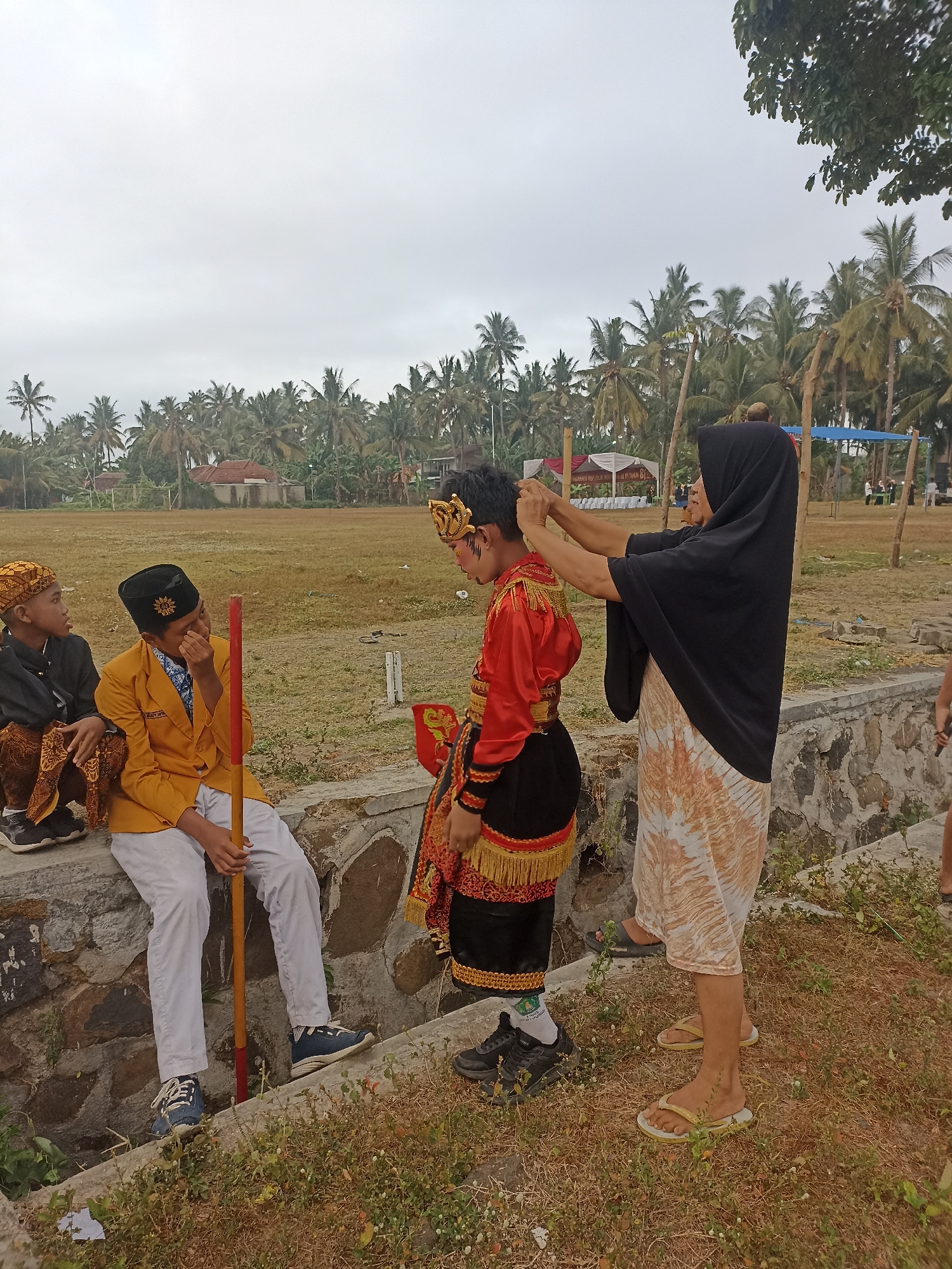 In Banyuwangi, Indonesia, an outdoor scene unfolds with a cloudy sky and a backdrop of numerous palm trees, indicative of a tropical coastal environment. Three young males are present, two of whom are seated on a low stone wall. One boy on the left wears a patterned headscarf and traditional clothing with a brown sarong. The boy next to him, in the center, is dressed in a bright yellow jacket over a blue and white patterned shirt, white pants, and blue sneakers. He holds a red and yellow pole. The third boy, positioned in the center-right of the frame, stands facing away from the viewer and is adorned in a red, gold, and black traditional outfit, complete with a golden headdress. A woman, wearing a black hijab and a patterned dress, stands behind the boy in traditional attire, adjusting his headdress. The ground is covered with dry grass and scattered weeds. In the background, a field is visible with some structures and tents, suggesting an event or gathering is taking place. The overall atmosphere appears to be preparatory, with the woman attending to the costumed boy's appearance.