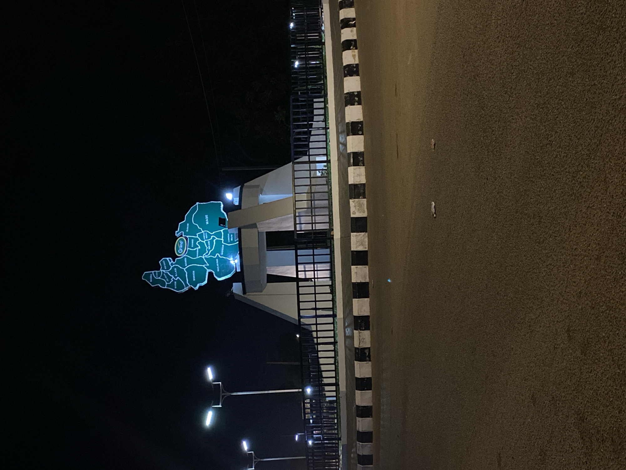 The image displays a nighttime outdoor scene in Bukuru, Nigeria, centered on an illuminated monument next to a road. The main subject is a large, glowing blue-green sign or monument shaped like a geographical map, likely depicting Plateau State, Nigeria, and its local government areas. Several names are visible on the map, including "WASE," "KANKE," "MIKANG," "QUAN PAN," "GULAK PAN," "BASIN," "OYON," "K.LAFI," "DOKKOS," and "BOKKOS." The word "Grand" is also visible on the map.
The monument stands on a base adjacent to a modern-looking structure or building, partially obscured in the background. A dark metal fence or railing runs along the side of this structure, separating it from the road. The asphalt road features a clear black and white striped curb or barrier. The scene is well-lit by several modern streetlights with bright, rectangular fixtures, indicating it is nighttime. The sky is dark, and no stars or moon are visible. There are no people or vehicles present, suggesting a quiet moment on what appears to be a public thoroughfare. The weather appears clear.