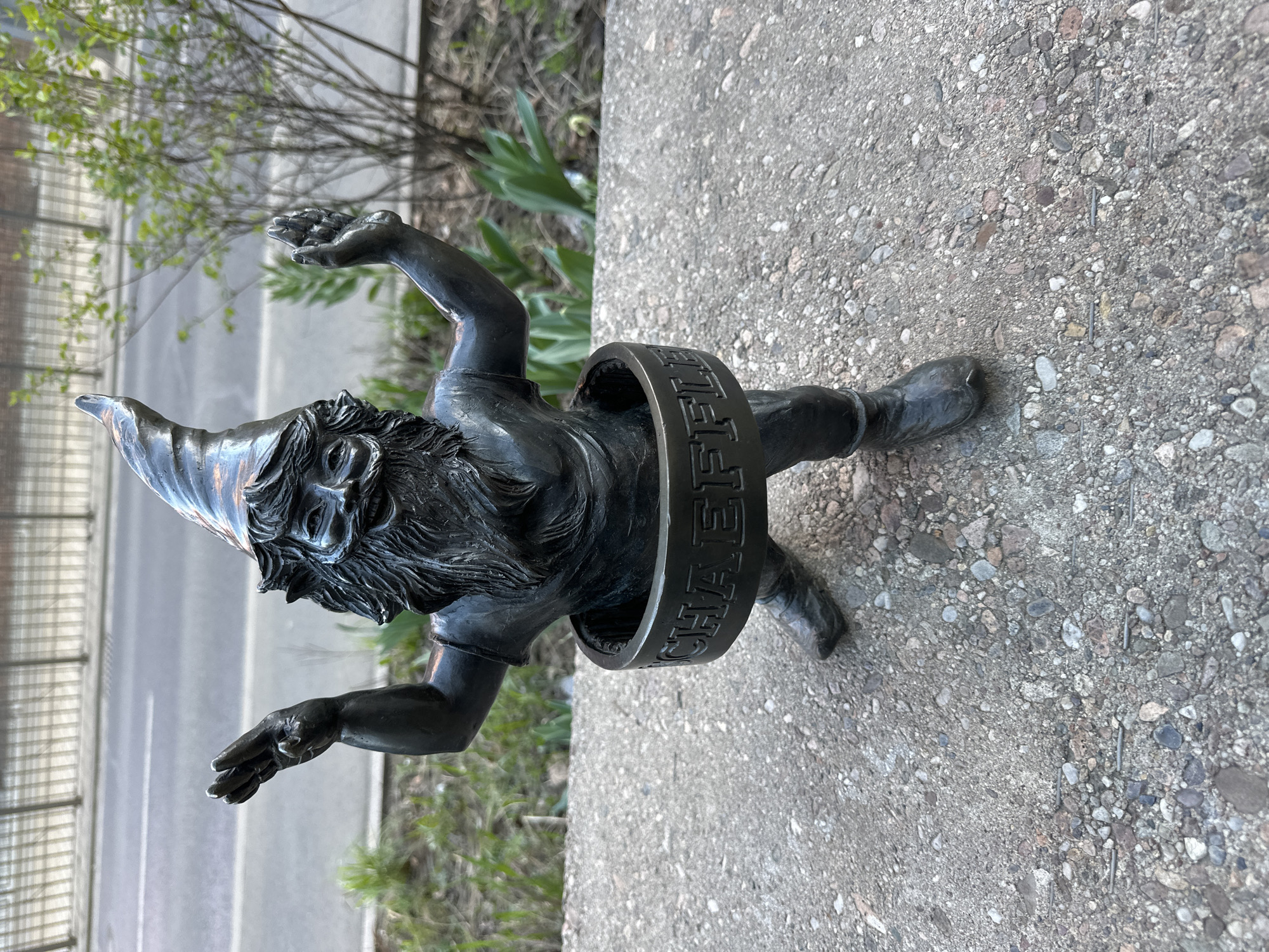 The image shows a bronze dwarf statue, known as a "Wrocław dwarf", in Wrocław, Poland. The dwarf is positioned on a gray concrete surface, likely a sidewalk or pavement. The statue is depicted in mid-action, with arms raised and legs straddling a metal ring. The ring has the inscription "MICHAEL SCHAEFFLER" in capital letters. The statue is detailed with a pointed hat, a beard, and facial features. The background includes greenery (bushes, trees), a roadway, and a building. The photo is taken at ground level with a slightly tilted perspective.