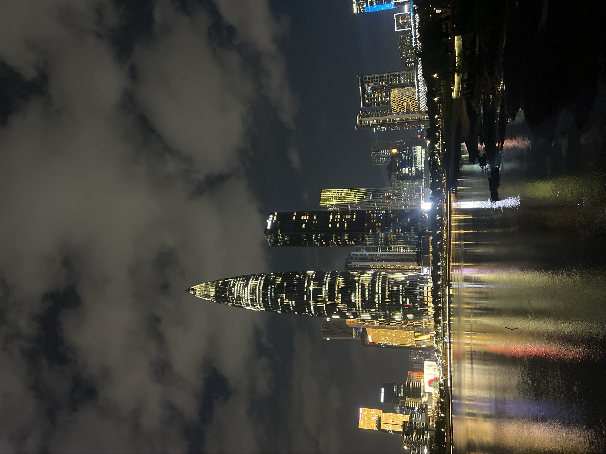 An atmospheric, nighttime, horizontal shot captures the illuminated skyline of Nanshan District, China, reflected in the calm waters of a bay. The iconic Ping An Finance Centre, a needle-like skyscraper, dominates the center of the frame, its windows glowing with warm light against a dark, cloudy sky. To its left, a cluster of shorter, brightly lit buildings with golden facades stand out. To the right of the Ping An Finance Centre, other skyscrapers of varying heights and architectural styles, also illuminated, form part of the cityscape. The water in the foreground mirrors the city lights, creating a vibrant, colorful reflection that stretches across the bottom half of the image. The shoreline is dimly visible, with trees and a pathway softly lit by streetlights. The overall impression is one of a modern, dynamic urban environment at night.