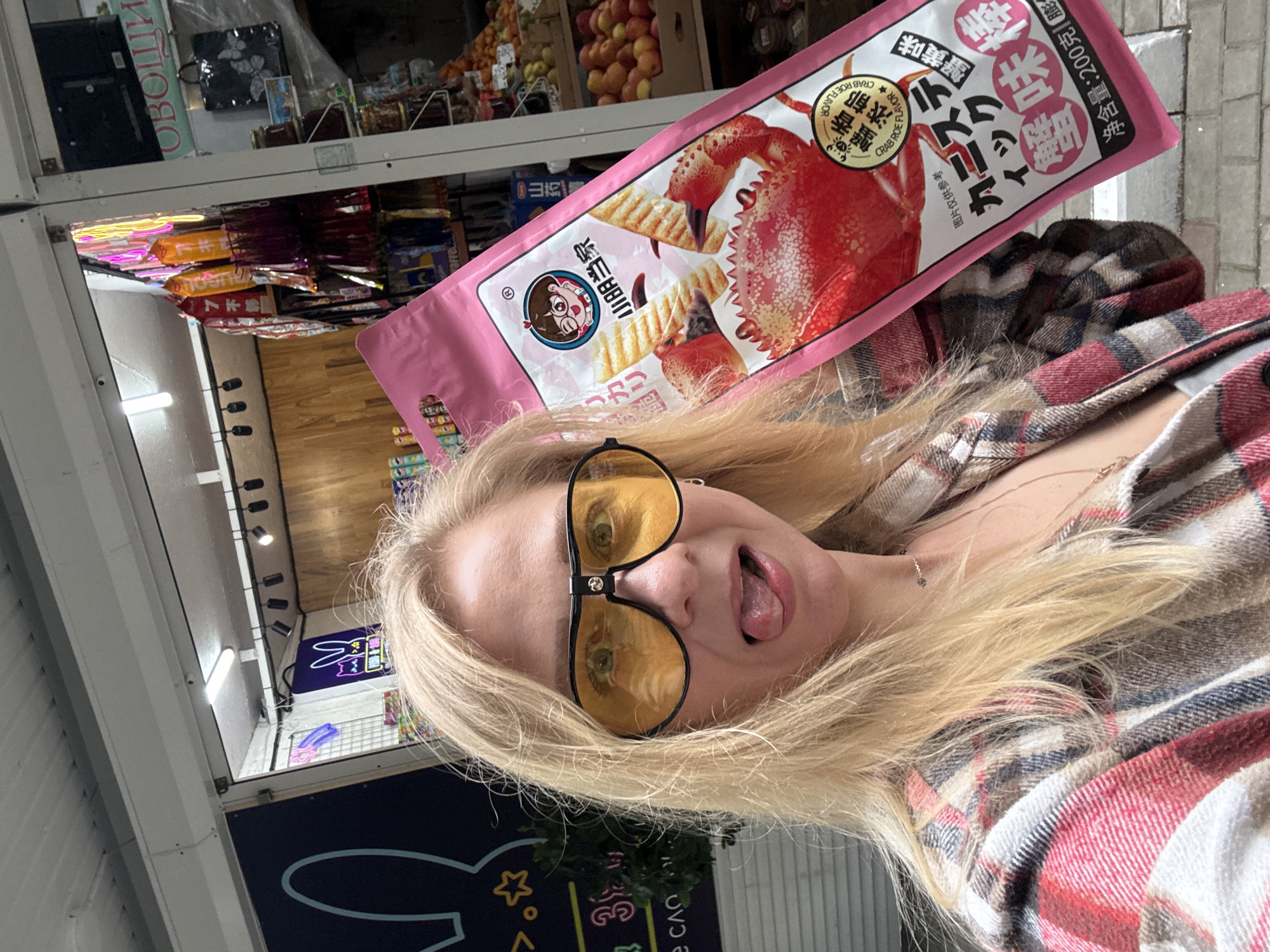 A blonde woman wearing yellow sunglasses is holding a pink package of crab-flavored chips. The package has an image of a crab claw on it. The woman is sticking her tongue out. Behind her is a storefront with a neon sign of a rabbit. There are also shelves of fruits and vegetables visible in the background.
