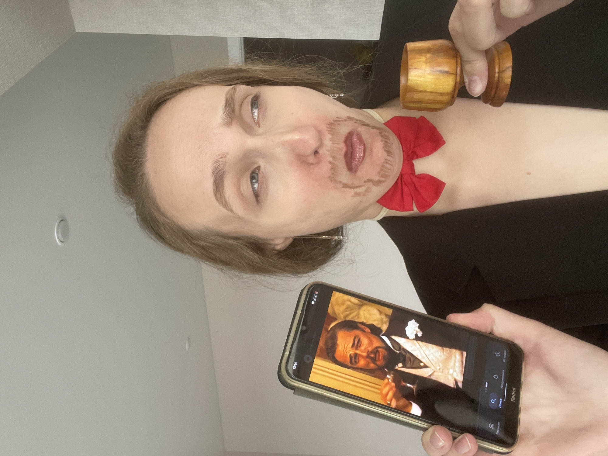 A woman lies on her side, holding a phone in her right hand showing a video of a man in a tuxedo. She has a drawn-on mustache and beard, and a red bow tie is placed at her throat. Her left hand holds a wooden object, possibly a cup or container. The background appears to be an interior space with a light-colored wall. There is no information to indicate the location as Podshivalovo, Russia.