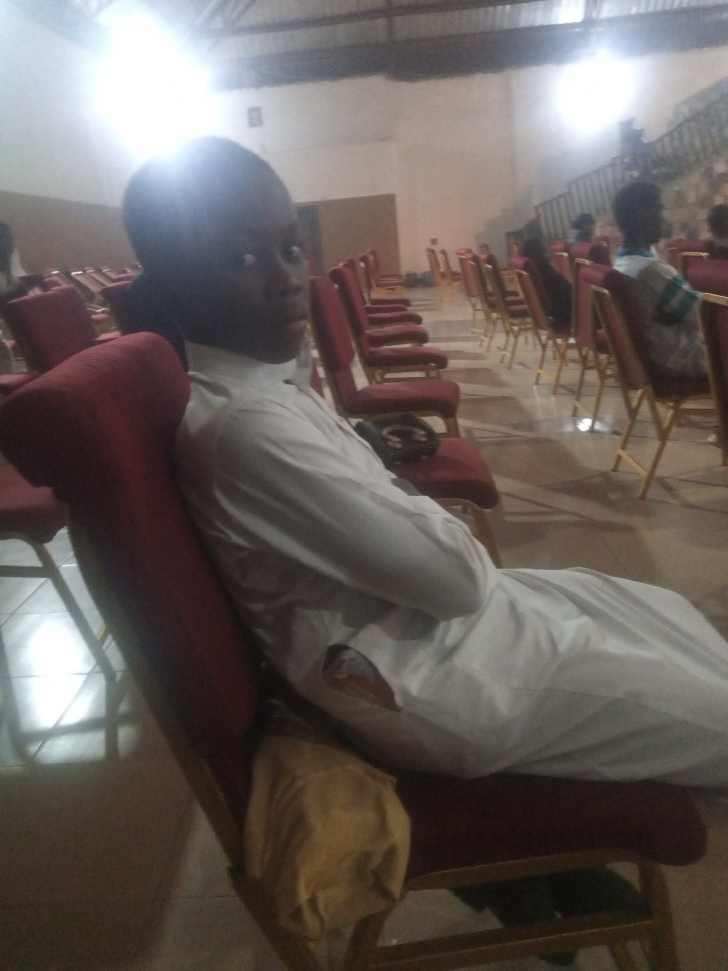 A young Black male, wearing a white tunic, is seated on a red upholstered chair in what appears to be an indoor event space. The camera angle is close-up, focusing on the male and the chair he occupies, with the background blurred and out of focus. Rows of similar red chairs stretch out behind him, suggesting an audience or gathering. The lighting is bright, with a strong light source in the upper left of the frame casting a glare. The floor appears to be tiled. The overall scene suggests a casual indoor event or service. This image was taken in Rukuba Road, Nigeria.
