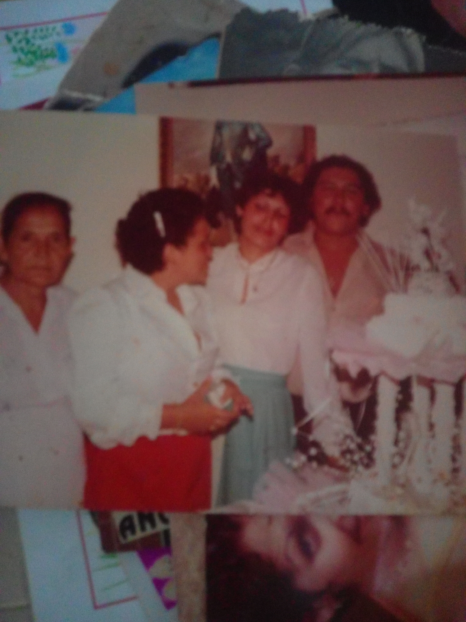 This is a blurry, waist-up photograph of four adults posing for a picture, likely at a celebration. On the far left is a woman with dark hair, facing forward with her head slightly tilted. Next to her is another woman, also with dark hair pulled back, wearing a white top and a red skirt, and she is looking to her right. To the right of her is a man with dark hair and a mustache, wearing a light-colored open-necked shirt. He is looking towards the right. Partially visible behind him is a fourth person, a woman in a white top, also looking to the right. The background appears to be a wall, and there is a large, white, tiered object in the foreground, possibly a cake stand with a cake. The lighting is soft and diffused. The photograph is part of a larger image, with the bottom portion showing a colorful design and text. There is no information about the location being La Tagua, Venezuela within the image itself.