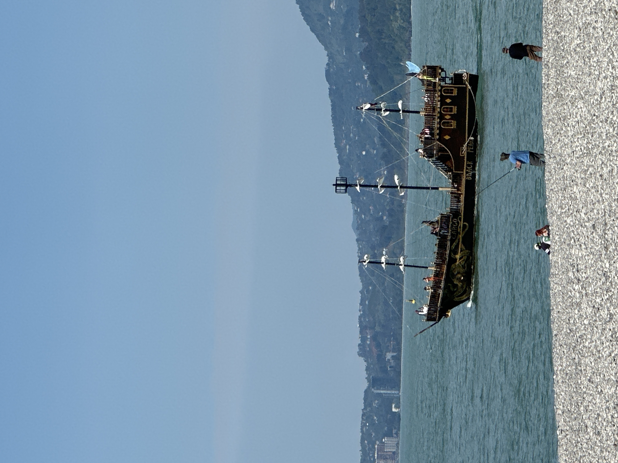 Here is a forensic description of the image:
The image depicts a large, elaborate pirate-themed ship anchored near a stony beach on a clear day. The ship, named "Black Pearl" and "Argo," has multiple masts and detailed decorations. Several people are standing on the beach next to the ship. The backdrop shows a mountain range and a coastline with buildings, consistent with the city of Batumi, Georgia. The overall environment is bright and sunny.
