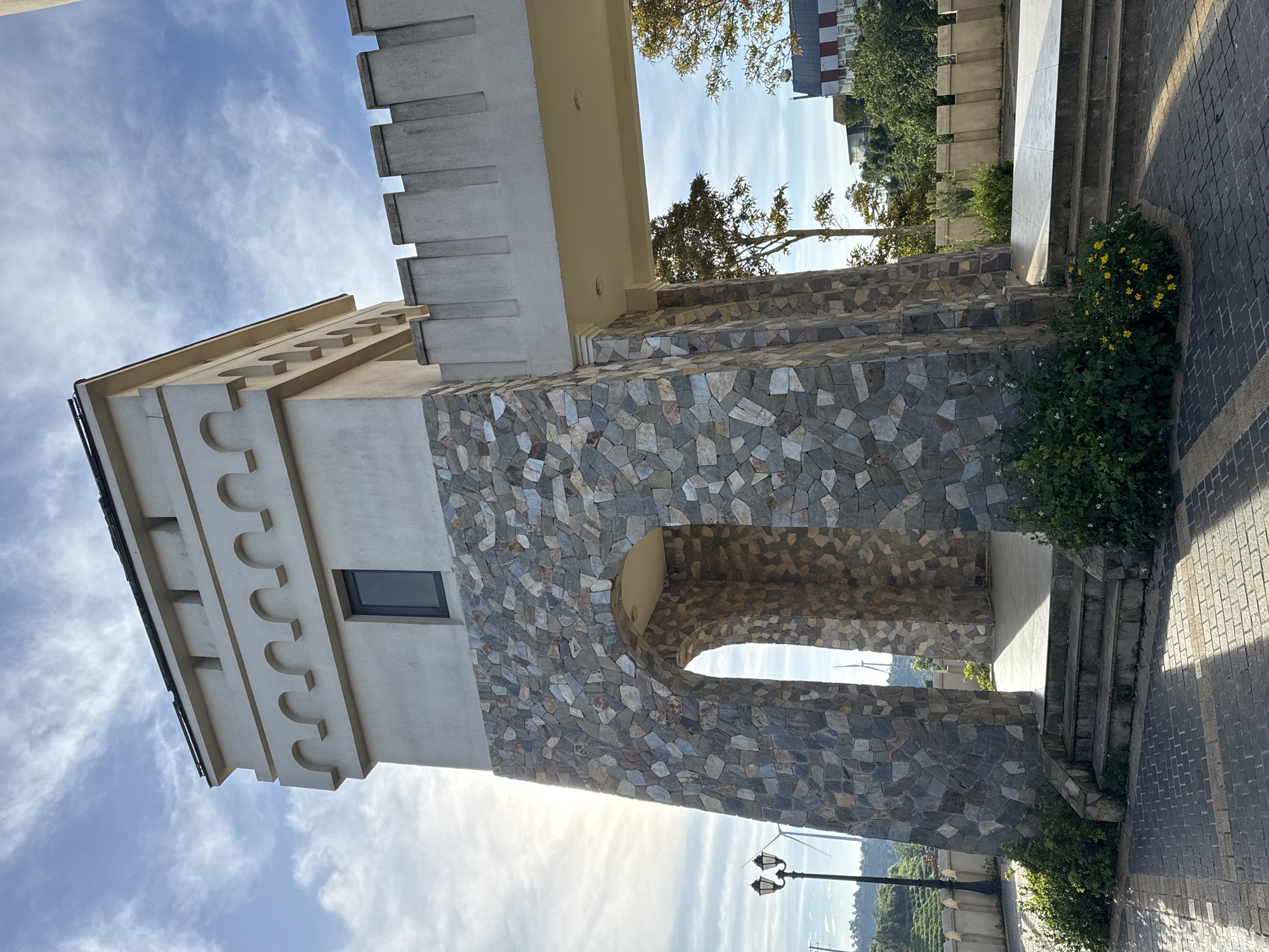 The image shows a stone tower with an archway, part of a building or complex designed in a castle-like style, located in Phát Chi, Vietnam. The tower has a light-colored upper section with decorative battlements and a dark rectangular window. The lower portion is covered in irregular grey, brown, and blue stone tiles. A paved walkway with a brick pattern leads to a set of stone stairs ascending to the archway. Green bushes and a few yellow flowers are visible around the base of the tower. To the left, a lamppost with two lights stands near a fence. In the background, trees, other buildings with red and white accents, and windmills are visible under a partly cloudy sky with sunlight streaming from the left. The scene appears to be an outdoor public space or entrance.