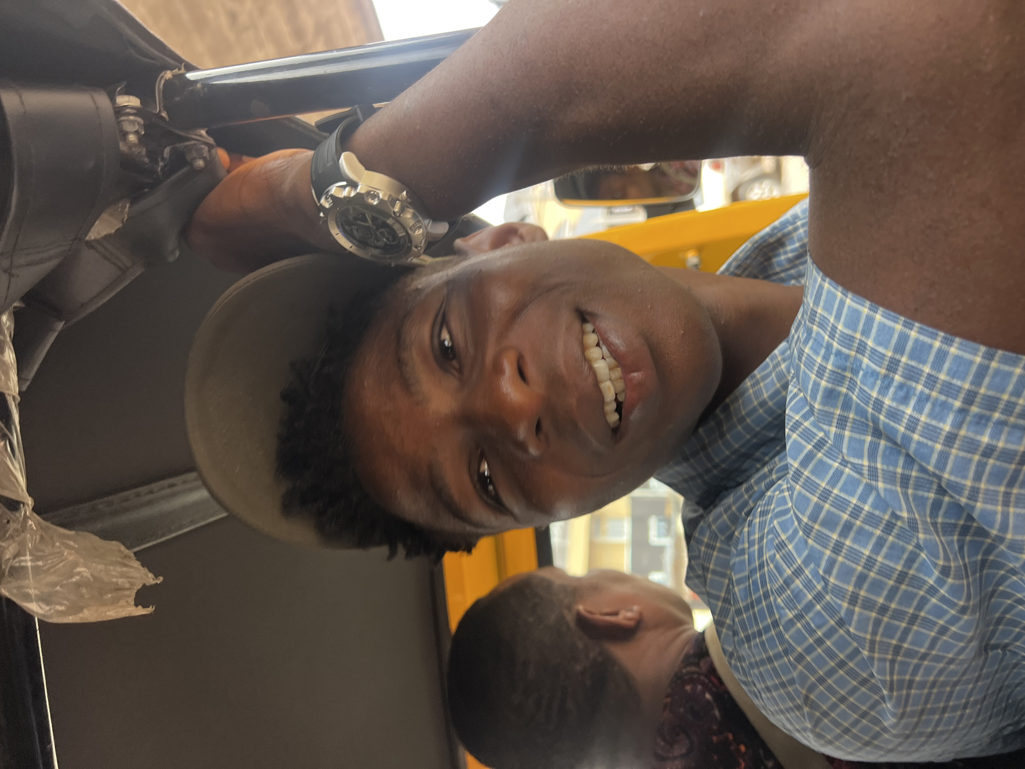 A close-up, front-facing shot captures a young Black man smiling while riding in a motorized tricycle, commonly known as an "okada" or "keke napep," in Nkpor, Nigeria. He is wearing a blue and white checkered shirt and a light-colored cap. His left arm rests on the tricycle's roof support, and he is wearing a large, silver-faced wristwatch on his left wrist. The background shows another passenger in the tricycle and glimpses of the urban environment outside, including buildings and vehicles. The lighting suggests it is daytime, with some harsh shadows.