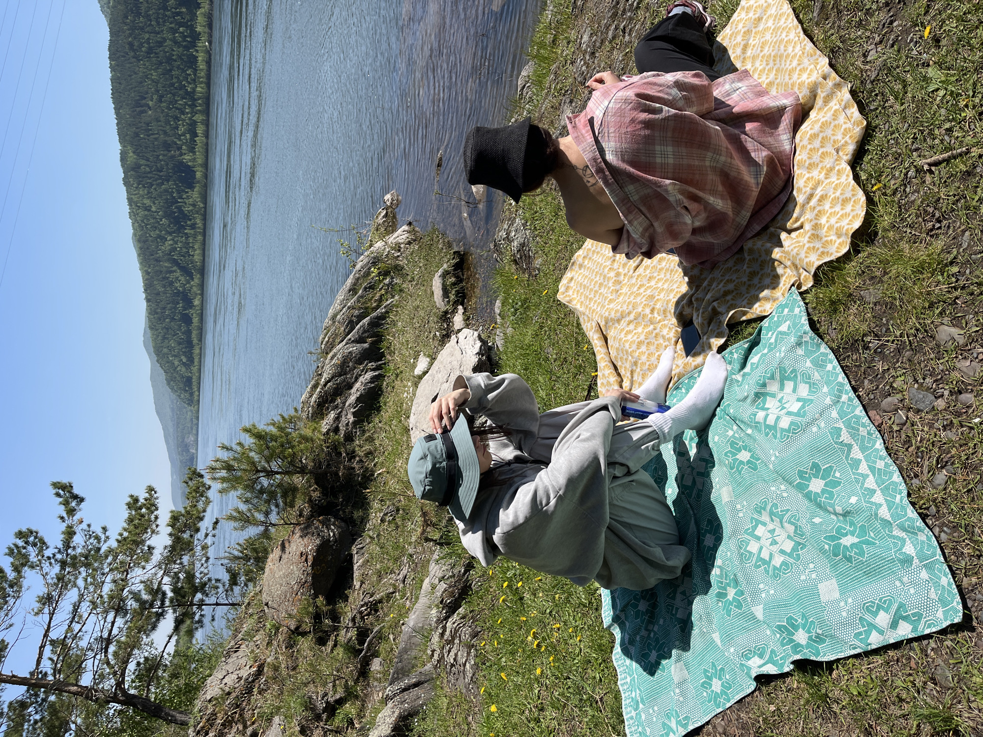 The image captures two individuals relaxing on the grassy bank of a wide river or lake on a bright, sunny day. The scene is set outdoors, with a backdrop of a dense, green forest covering distant hills or mountains on the opposite side of the water. The clear sky and strong shadows indicate midday or early afternoon.
In the foreground, to the left, one person is dressed in a grey hooded sweatshirt and pants, a light blue baseball cap, and white socks. They are seated on a teal blanket adorned with white geometric patterns and appear to be looking down at a white object held in their hands, possibly a phone or book, with some partially obscured text visible.
To the right, the second person wears a red and white plaid shirt, black pants, and a black bucket hat. They are seated on a yellow blanket with a subtle repeating pattern and face away from the camera, looking towards the water or slightly to the side. A tattoo is visible on their right arm. A black smartphone rests on the yellow blanket near them.
The immediate riverbank is rugged, featuring rocks, patches of green grass, and small trees. The water surface shows slight ripples, reflecting the clear blue sky. The overall impression is one of peaceful outdoor relaxation in a natural environment.