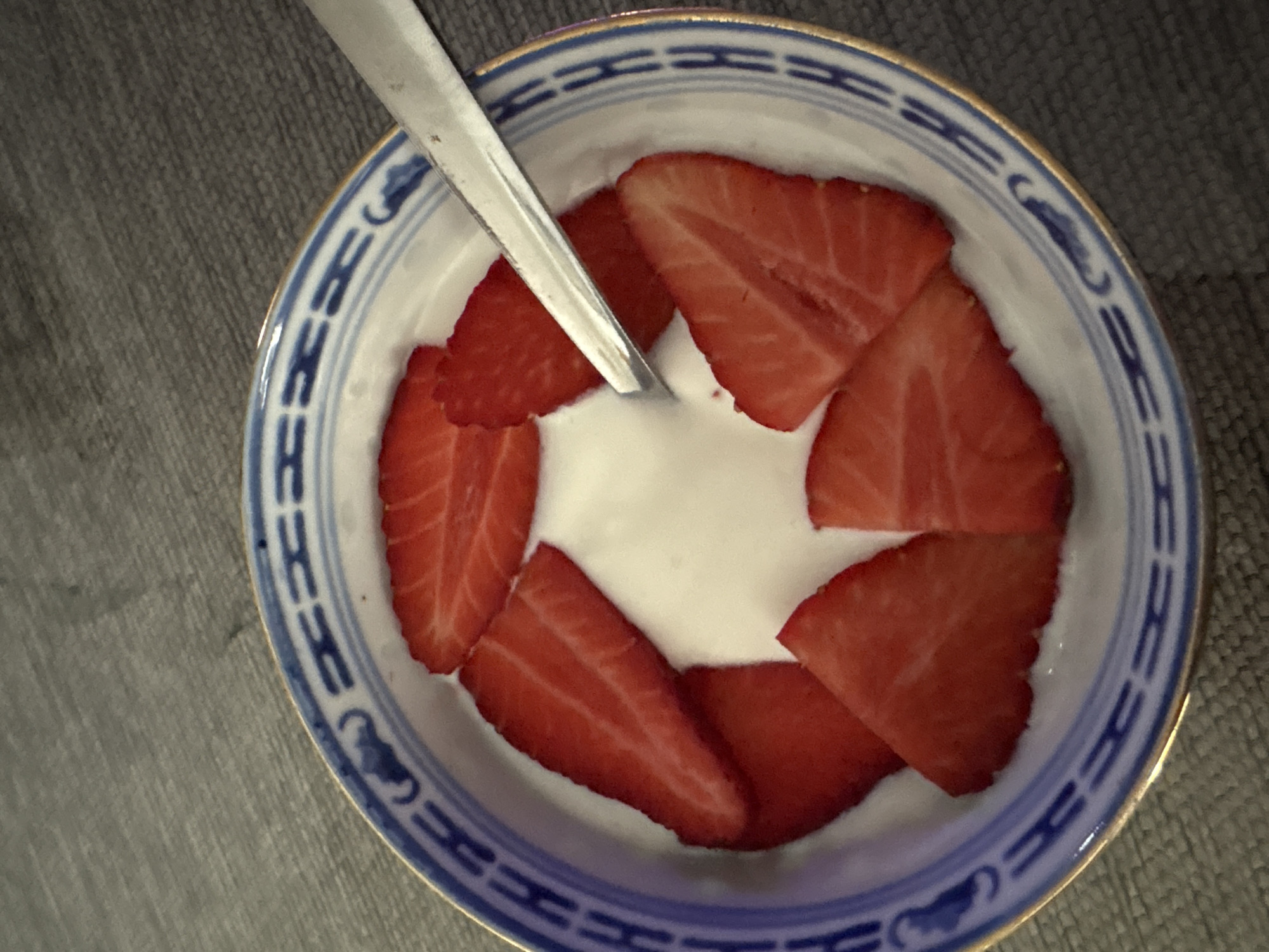 A top-down, close-up shot features a white ceramic bowl with a blue and gold decorative rim. Inside the bowl, slices of fresh strawberries are arranged around a white, creamy substance, likely yogurt. A silver spoon rests within the bowl, partially submerged in the yogurt. The background appears to be a gray, textured surface. The image was taken at an undefined location within Urbanización Encinares De Alcolea, Spain.