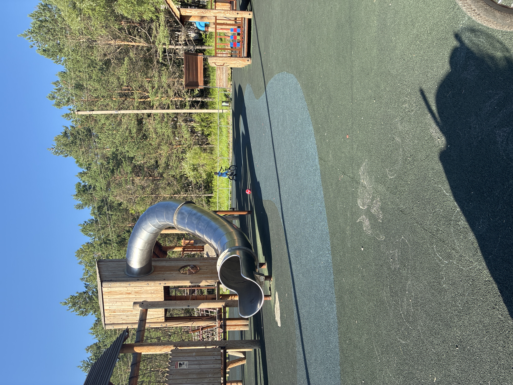 A child on a bicycle rides on a playground with a large metal slide and wooden structures. The playground has a green and blue rubberized surface. In the background, there are tall pine trees and a clear blue sky. The image is taken in the city of Gornye Kljuchi, Russia.