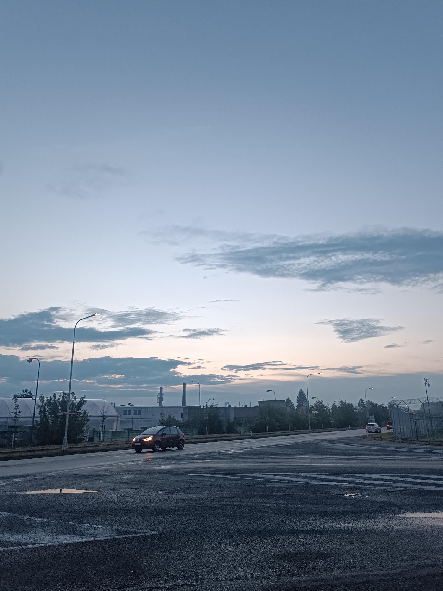 A wide-angle, low-light shot captures a desolate, wet intersection in Přelouč, Czechia, possibly at dawn or dusk. The asphalt road surface is slick with moisture, reflecting the faint light from the overcast sky and a single distant red traffic light. A dark red hatchback car is the only visible vehicle on the road, driving away from the viewer toward the right. The background features a long, low-rise industrial building complex with multiple smokestacks or towers, obscured by a line of trees. Streetlights with illuminated bulbs are positioned along the roadside. The sky is a pale blue gradient with scattered, wispy clouds, suggesting an early morning or late evening scene. The overall impression is one of a quiet, functional industrial area on a damp day.