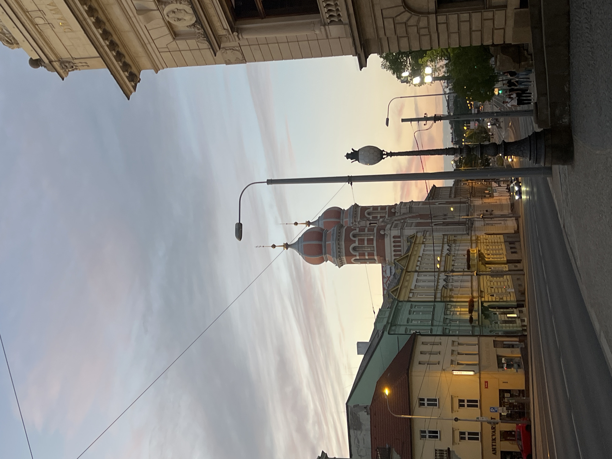 The image shows a street scene in Pilsen, Czechia, at dusk. The sky is a soft blend of pink, orange, and grey clouds, indicating the end of the day. In the foreground, a paved sidewalk runs along a street with asphalt pavement. A tall, ornate lamppost with a decorative base and globe-shaped light fixture stands prominently on the sidewalk. Further down the street, a modern lamppost is visible.
On the left side of the street, a row of colorful buildings lines the road. The closest building is yellow with an "ANTIQUARY P" sign visible on its facade. Next to it is a light green building, followed by a yellow building with decorative elements. Across the street, a striking red and white building with two onion-shaped domes is the focal point. This building appears to be a significant architectural landmark.
On the right side of the street, a large, light-colored building with stone cladding and decorative architectural details dominates the view. The word "DRAMA" is visible in relief on its facade, suggesting it might be a theater or cultural building. Several people are walking on the sidewalk in front of this building.
Vehicles are present on the street, including a red car parked on the left and other cars driving on the road. Streetlights are illuminated, casting a warm glow on the surroundings. The overall impression is a picturesque urban landscape bathed in the soft light of twilight.