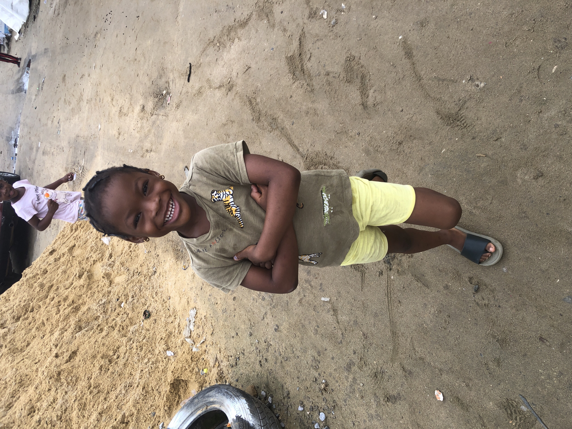 In the city of Ado, Nigeria, a young girl stands with her arms crossed, smiling at the camera. She is wearing a light brown t-shirt with a tiger design and bright yellow shorts. The girl has dark skin, braided hair, and gold hoop earrings. She is standing on a dirt and sand ground, which is littered with debris, including a discarded tire to her left. In the background, a pile of sand can be seen, and another young girl in a pink t-shirt and white shorts walks away from the camera. The scene appears to be an outdoor area, possibly a construction site or a dusty urban environment.