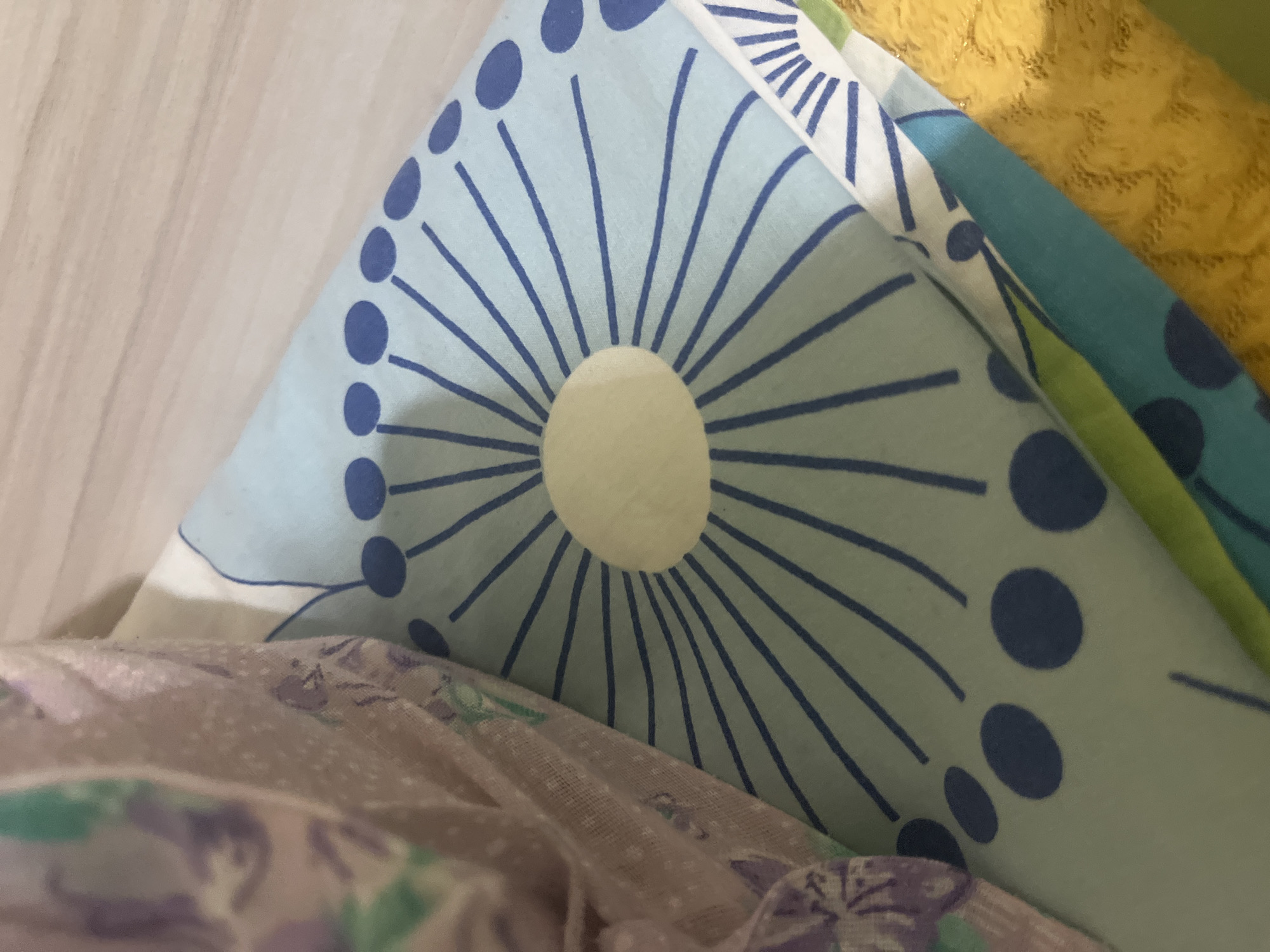 The image shows a close-up view of folded bedding with a patterned design. The dominant pattern features a light blue background with radial lines emanating from a pale yellow circular center, resembling a stylized sunburst or dandelion seed head. Dark blue dots are also present along some of the radial lines. A portion of a different fabric, a light purple material with faint floral or abstract patterns in purple and green, is visible on the left side of the image, draped over the blue patterned fabric. The bedding appears to be stacked or folded, with hints of other colored fabrics, including yellow and green, visible underneath and to the right. The background shows a light-colored, possibly wooden surface, suggesting the bedding is placed on a piece of furniture. The scene is likely indoors. There are no people or visible signs of activity. The lighting is soft, creating gentle shadows. The location in Burgas, Bulgaria is not discernible from the image itself.