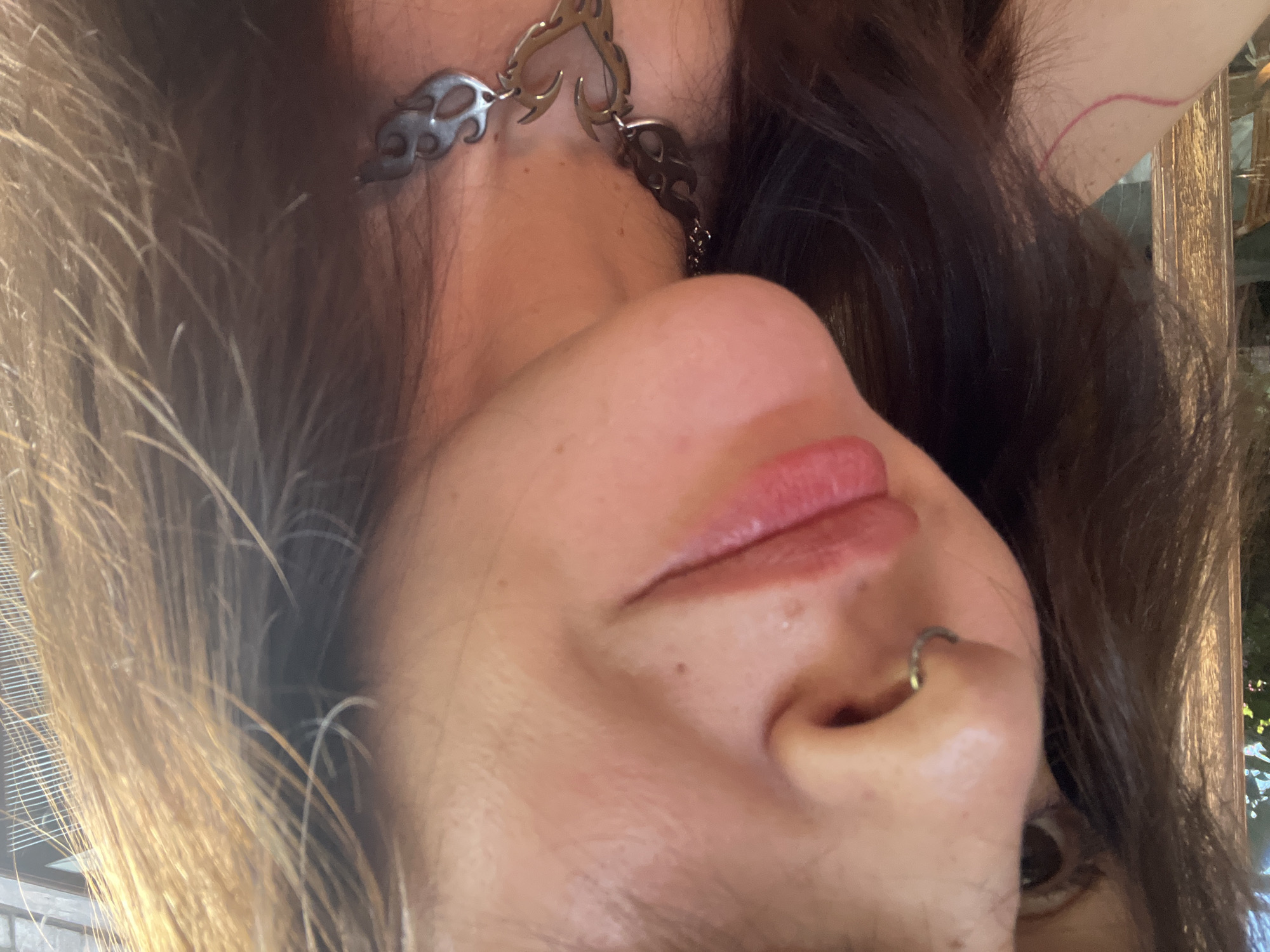 An upside-down selfie of a person with light skin, light brown and dark brown hair, and a nose ring. The person is wearing a metal necklace with flame-shaped links. The background is blurry, with hints of outdoor foliage and architectural elements. The location in the city of North Kuta, Indonesia, cannot be determined from the image.