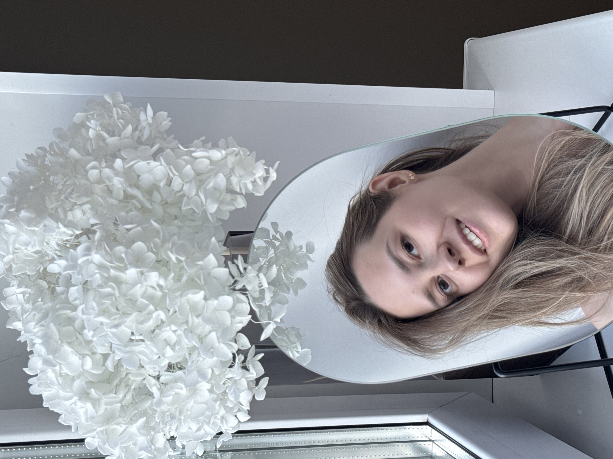 The image shows a person's reflection in a round mirror positioned on a white table, alongside a bouquet of white flowers. The reflection captures the person's face, with their head tilted. The setting appears to be indoors, with a window visible at the bottom of the frame. The background includes a neutral-colored wall and a glimpse of what seems to be a white chair. There is no information in the image to indicate that it was taken in Chaikovsky, Russia.