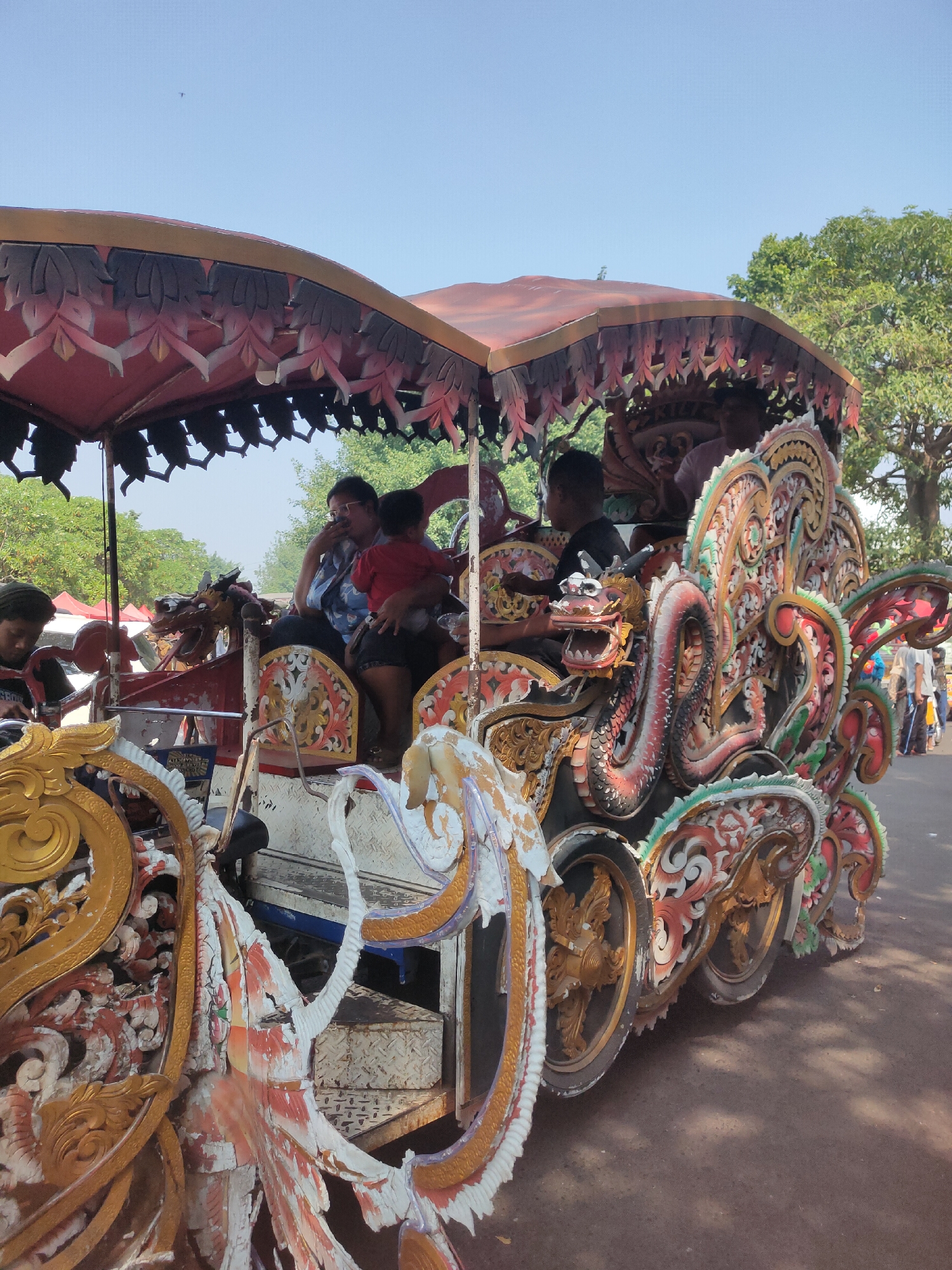 The image shows a brightly decorated horse-drawn carriage with people inside, likely in the city of Ngasem, Indonesia, given the cultural context suggested by the carriage's ornamentation. The carriage is intricately carved and painted with vibrant colors, featuring motifs that appear to be dragons and other mythical creatures. A canopy of red fabric with decorative trim covers the top of the carriage. Inside, several individuals are visible. In the foreground, a person in a blue shirt with glasses is holding a child dressed in red. Another person, wearing a dark shirt, is seated next to them. The carriage has ornate wheels and is positioned on a dirt or gravel road. In the background, trees, tents, and other people are visible, suggesting a public event or festival. The sky is clear and blue, indicating a sunny day.