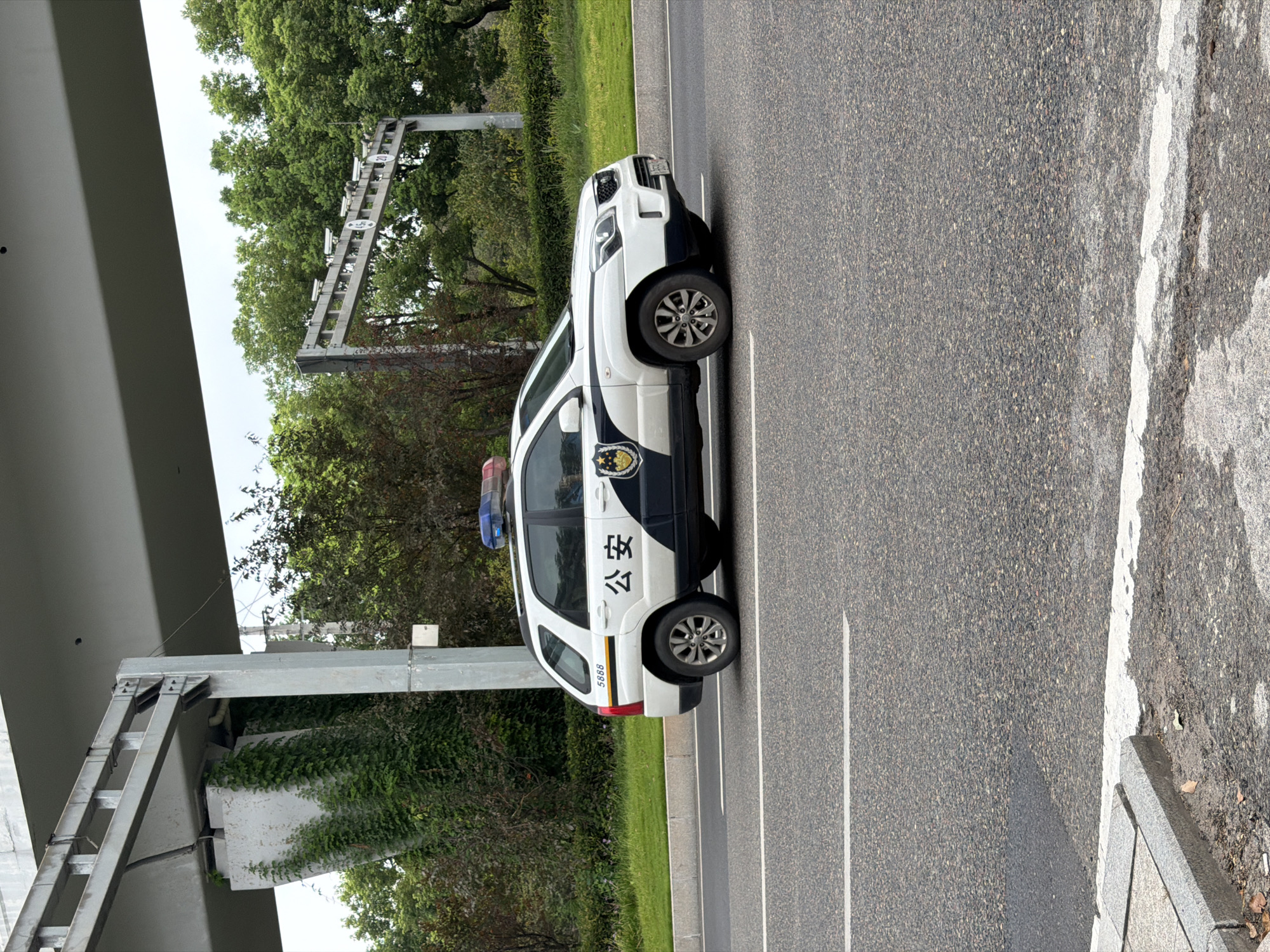 The image shows a white police SUV, identified with Chinese characters "公安" (Gōng'ān, meaning public security) and the number "5888," driving on a paved road. The vehicle has a light bar with red and blue lights on its roof. In the background, there is a large overpass structure, lush green trees, and a cloudy sky. Traffic cameras are visible mounted on a gantry above the road. The location is identified as Jiangjin, China. The scene appears to be a normal daytime urban or suburban environment.