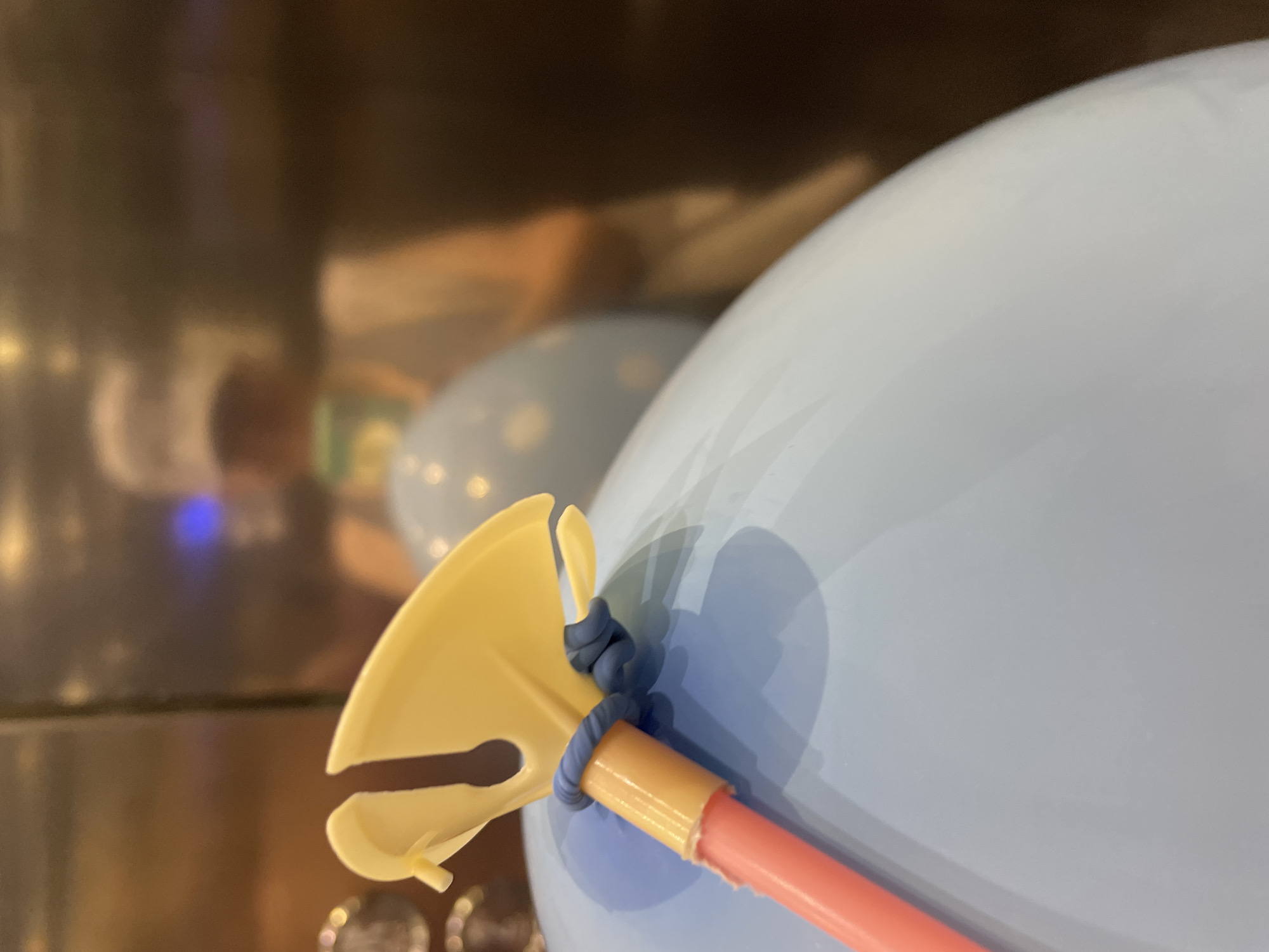 The image shows a close-up of a blue balloon with a yellow plastic balloon tying tool and a pink stick. The balloon is attached to the stick with a blue rubber band. In the background, there is a blurred reflection of a person wearing a white cap and a light-colored jacket, possibly in an elevator due to the metallic walls. The scene appears to be indoors. There are no discernible elements in the image that would indicate its location in Almaty, Kazakhstan.
