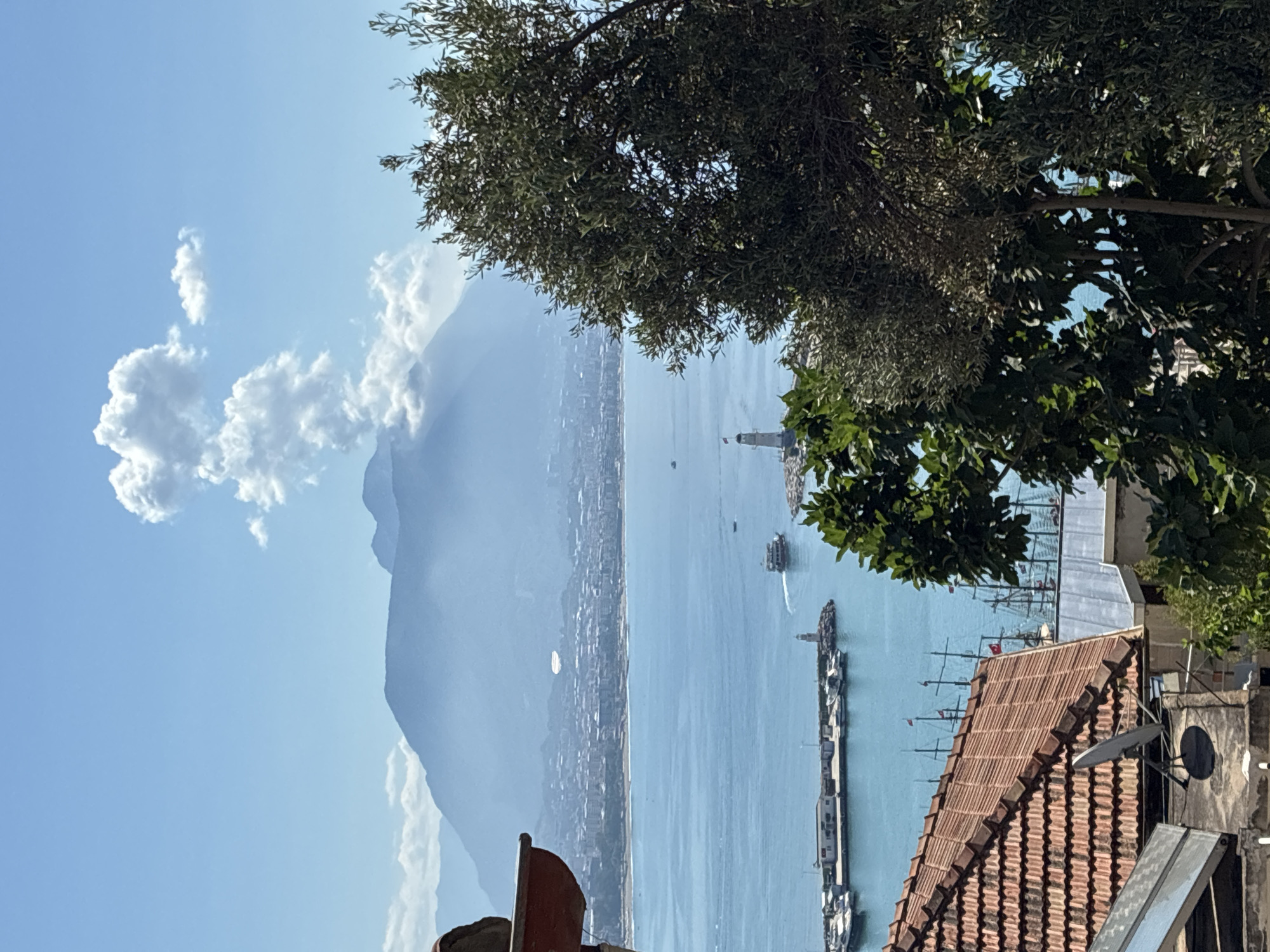 The image captures a scenic view of the Alanya coastline in Turkey, looking out over the Mediterranean Sea. In the foreground, a tiled red roof and a satellite dish are visible, suggesting a residential area. Beyond this, the vibrant blue sea stretches out, dotted with various boats, including several large sailing vessels with their masts.
A prominent lighthouse stands on a jetty, marking the entrance to a harbor filled with smaller boats. In the mid-ground, a city sprawls along the coastline, backed by a large, mist-covered mountain range. The sky is a clear blue, with a few fluffy white clouds scattered across it, one of which is dramatically positioned above the mountain peak. The overall impression is one of a sun-drenched day in a coastal city.