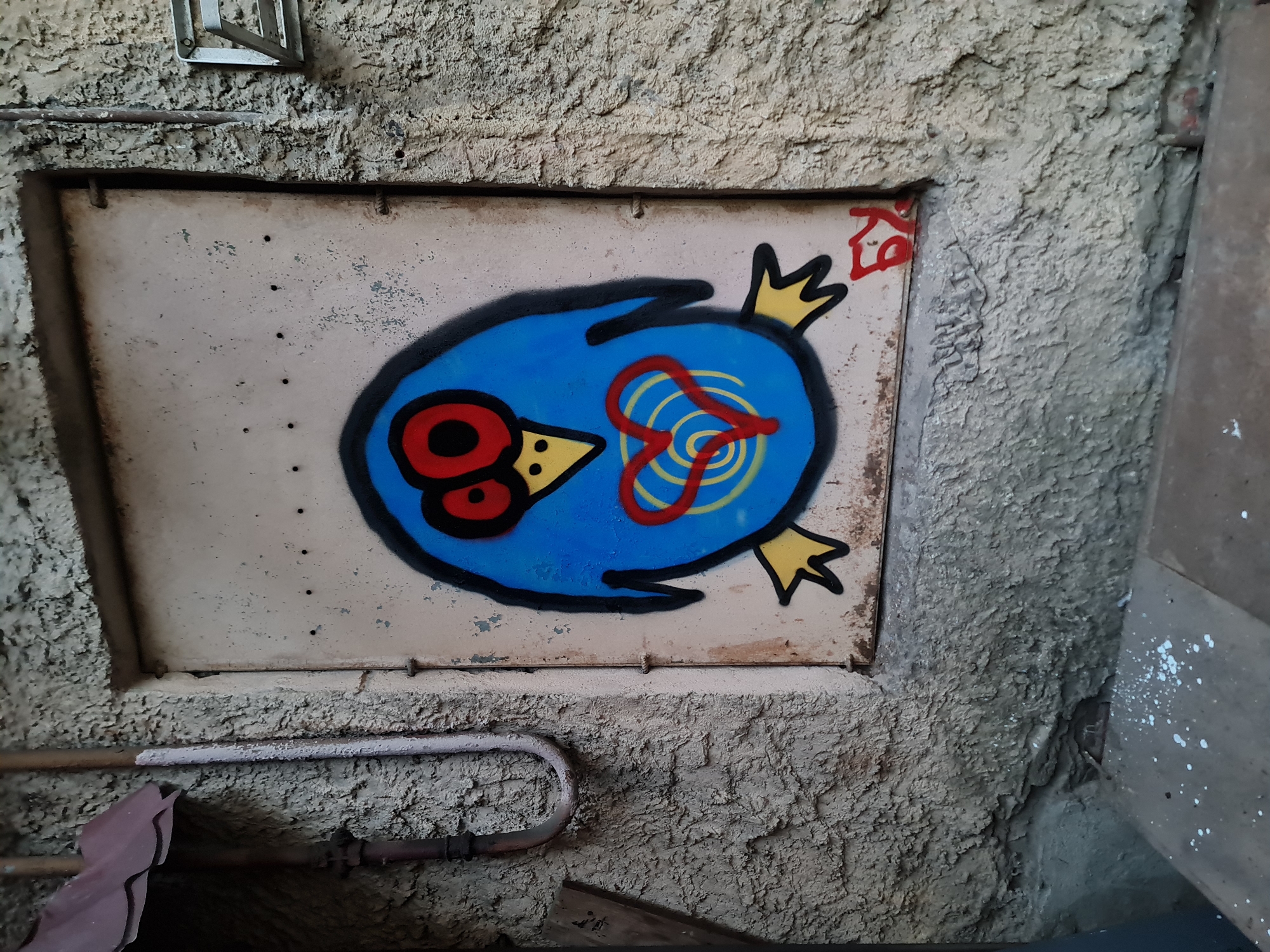 A graffiti of a blue bird with red eyes and a heart on its chest is spray-painted on a metal door. The door is set into a rough, textured concrete wall. To the left of the door, a metal pipe curves downwards, attached to a darker pipe. Above the door, a dark, ridged awning is visible. The scene is located in Saint Petersburg, Russia.