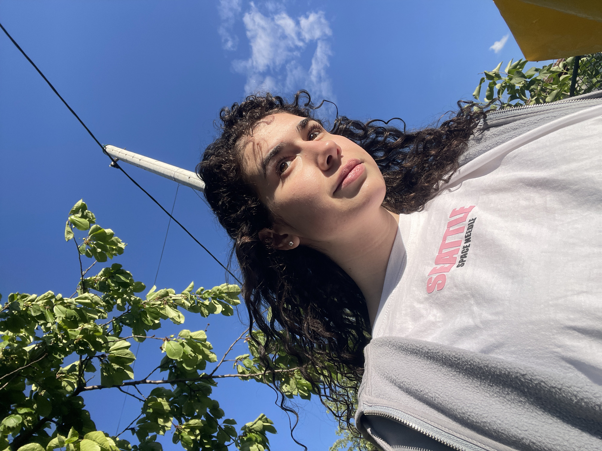 A low-angle, medium shot captures a young woman with curly dark hair, fair skin, and light makeup. She's wearing a white t-shirt with pink lettering ("SPACE NEEDLE") and a grey zip-up jacket. She is looking upwards and slightly to her left. The background consists of a bright blue sky with scattered clouds, green tree foliage on the left side of the frame, and a white pole with a black wire running parallel to it. The presence of the "SPACE NEEDLE" t-shirt doesn't provide enough information to determine a specific location in Sofia, Bulgaria. The rest of the scene does not offer additional forensic information.