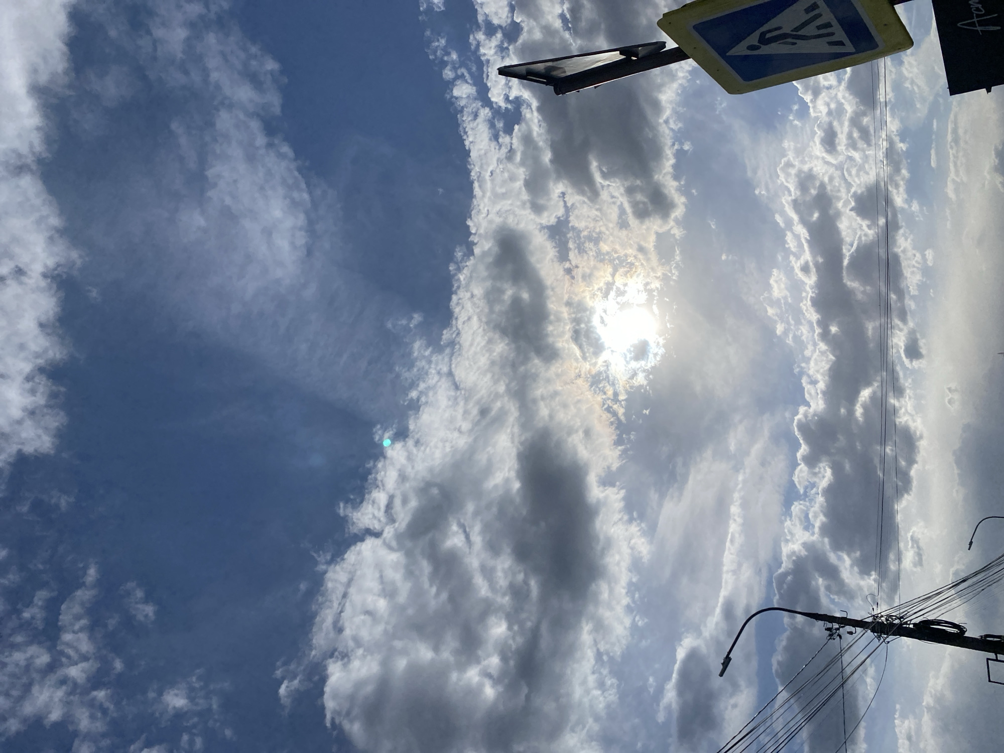 The image shows a sky view in Chaikovsky, Russia, with white and gray clouds and blue sky. The sun is shining brightly through the clouds. In the upper right, a pedestrian crossing sign is partially visible. Overhead electrical wires and a street lamp are visible in the lower right.