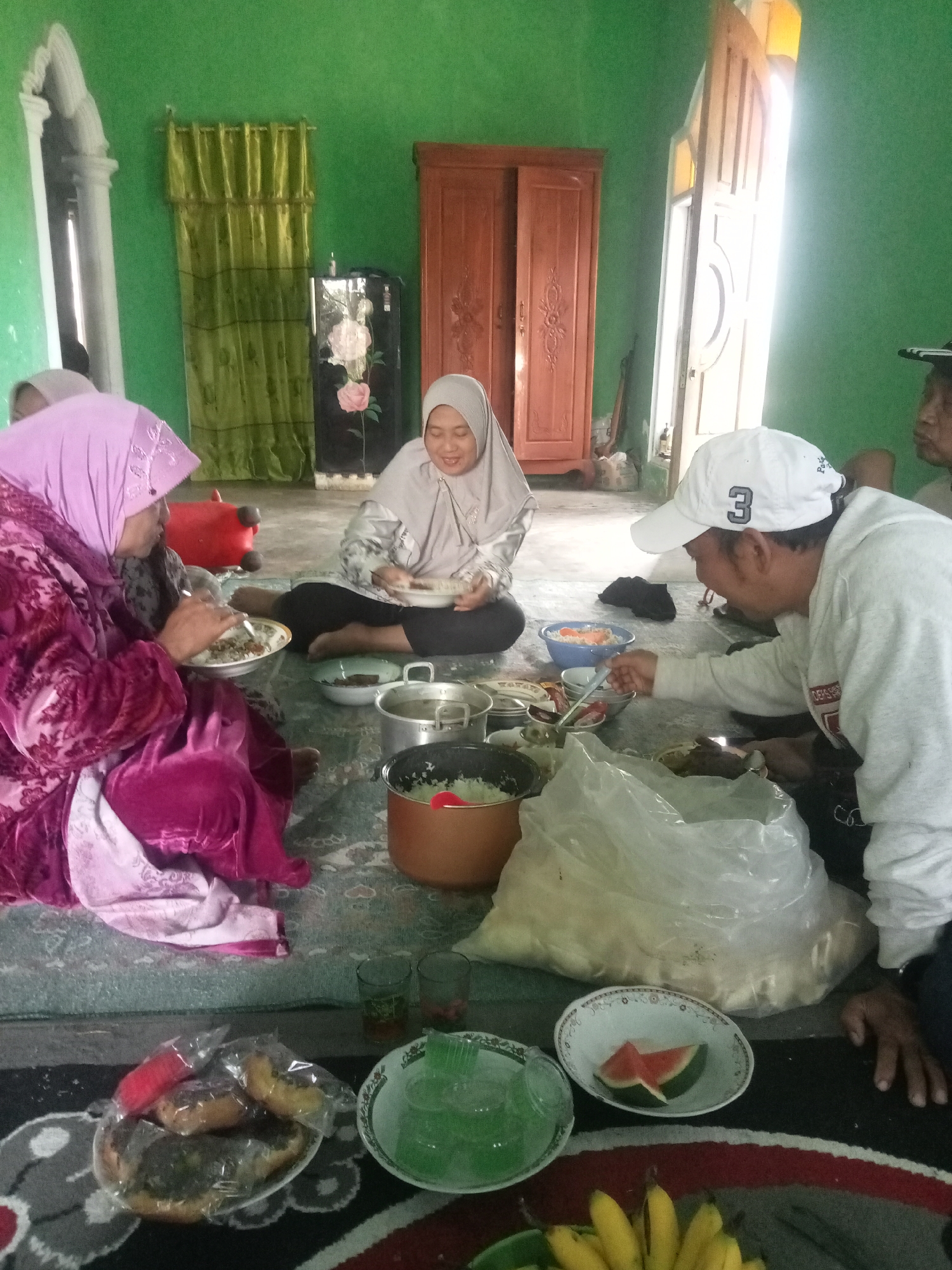 This image depicts a gathering of people in what appears to be a home in Tirtomoyo, Indonesia. Several individuals are seated on the floor, sharing a meal. One woman, wearing a pink headscarf and a vibrant pink garment, is actively eating from a bowl. Another woman, in a light-colored headscarf, is also eating. A man wearing a white baseball cap with the number "3" on it is reaching for food. The room is sparsely decorated, with a wooden wardrobe visible in the background. Green curtains hang near a doorway, and a large, white plastic bag, likely containing food, is present. Plates of food, including rice and what looks like fruit, are spread across the floor. The overall atmosphere suggests a casual, communal dining experience.