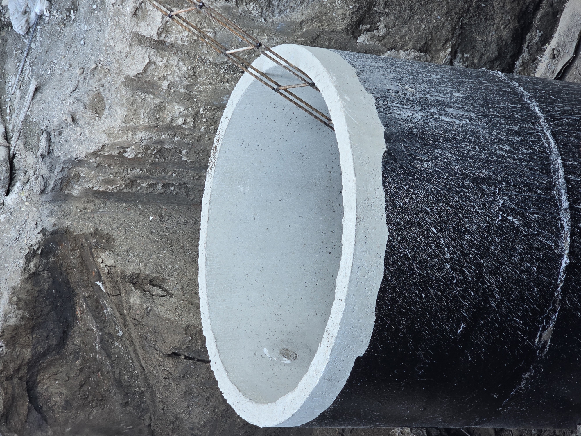 This is a medium-shot, close-up, eye-level, outdoor, daytime photograph of a large, concrete pipe section at a construction site in Perugia, Italy. The pipe is black on the bottom half and white on the top half, with a beveled edge where the colors meet. The interior of the pipe is visible and appears to be made of concrete. Inside the pipe, a section of metal rebar cage is visible. The pipe is situated in a dug-out area with dark soil and rocks surrounding it. The soil shows signs of excavation and has a rough, uneven texture. The lighting is natural and casts soft shadows. The image is in focus and the colors are natural. There are no people visible in the image. The overall impression is one of ongoing construction work.