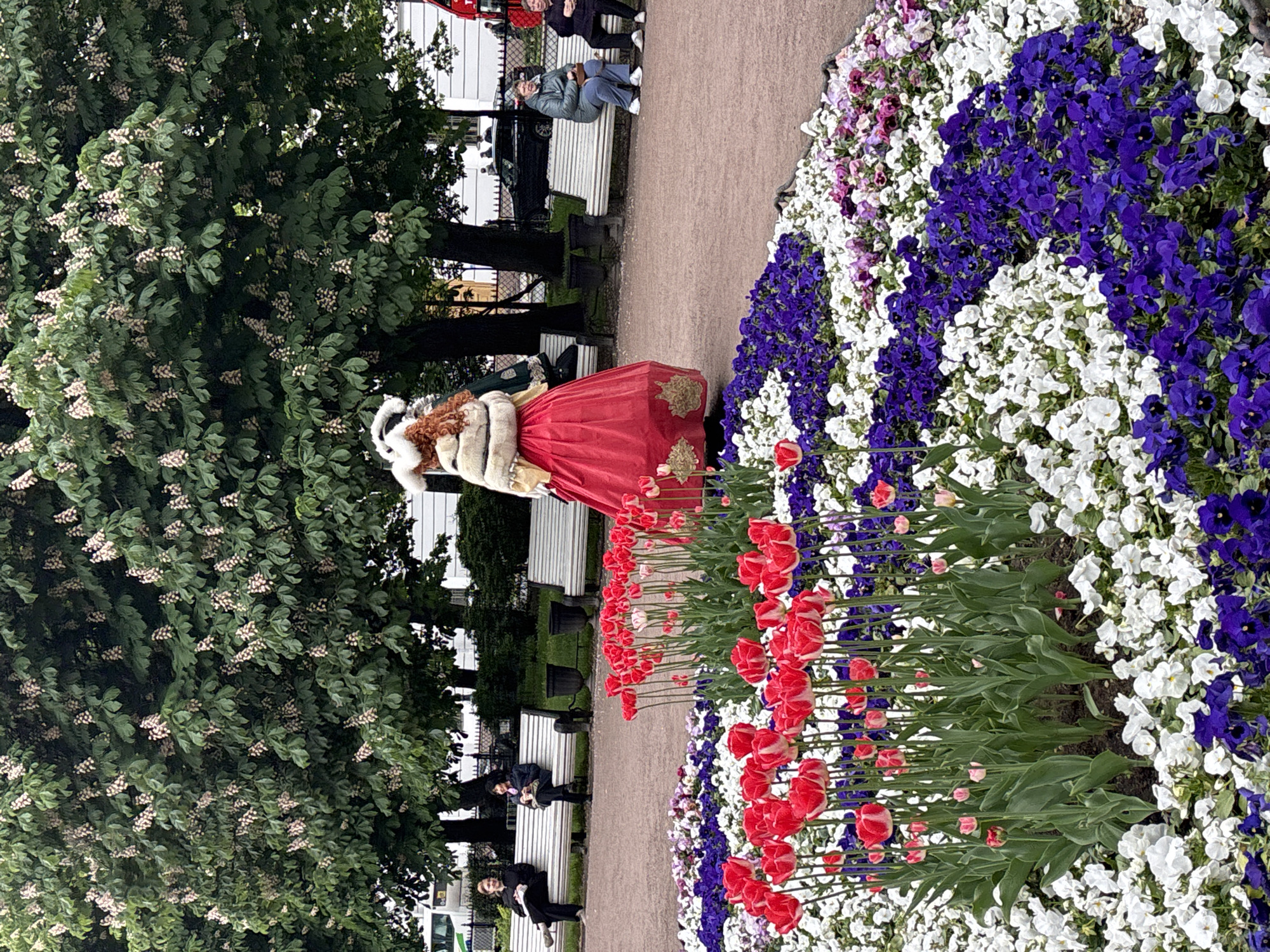 In Saint Petersburg, Russia, a woman in a red dress with a white fur-trimmed coat and hat stands beside a flower bed with white and purple flowers and red tulips. She faces left, looking toward the left side of the frame. Behind her, there is a path with benches on either side, on which a few people are seated. Trees with white flowers are also visible.