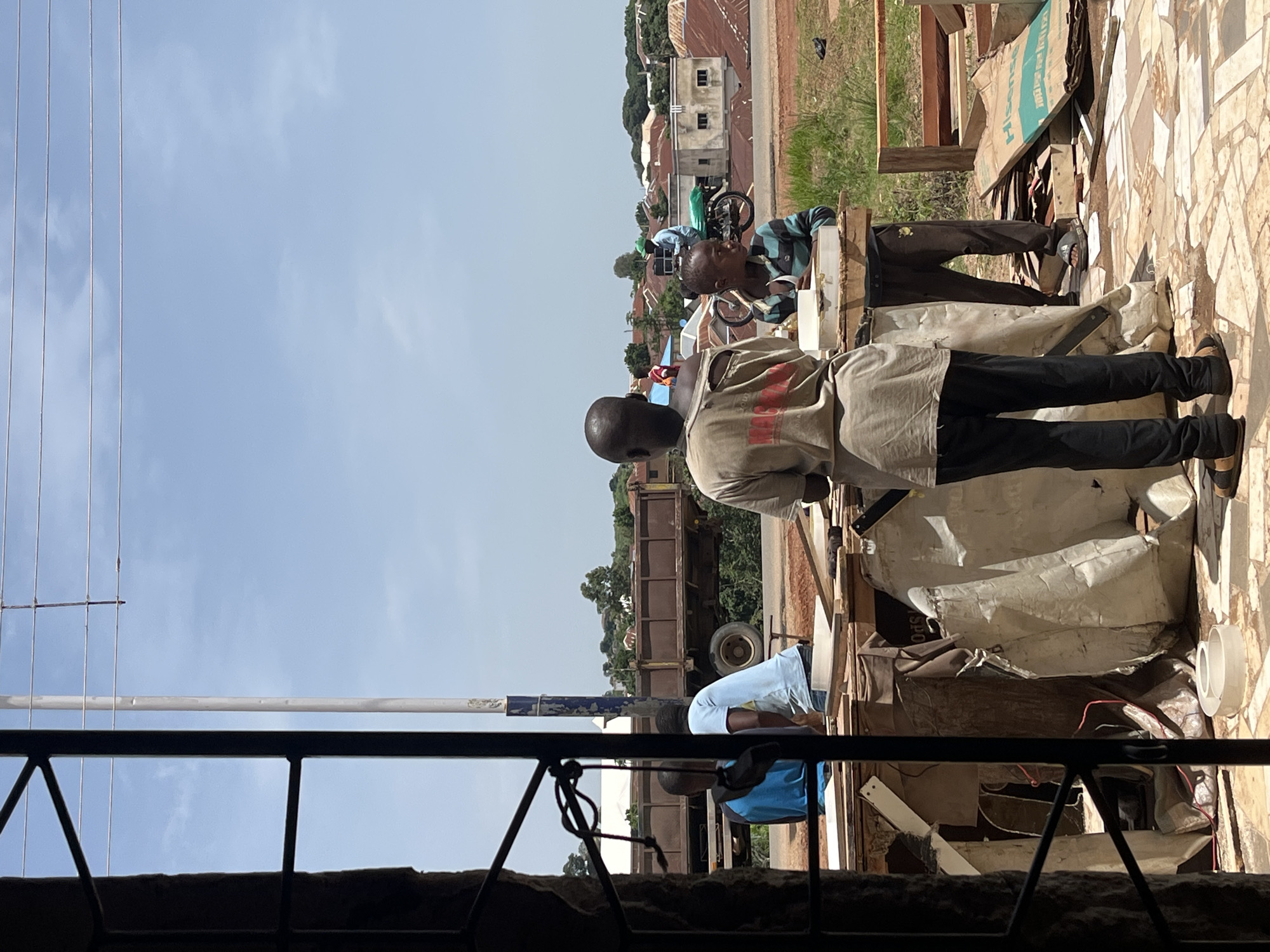 The image depicts an outdoor scene in Jos, Nigeria, with a focus on a group of young people in a workshop or construction area. The foreground is dominated by a structure with dark, vertical bars, suggesting a window or doorway through which the scene is viewed. Several young boys are the central figures. One boy, wearing a light-colored, torn t-shirt and black pants, stands with his back to the camera, looking towards the activity. Another boy in a striped shirt and dark pants is seated or leaning, appearing to work on something in front of him. A third boy in a blue shirt is also visible, engaged in some task.
In the background, a large, brown truck with a raised bed is parked on a dirt road. Beyond the truck, a cluster of buildings with red-tiled roofs signifies a residential or commercial area. The sky is clear and blue with some scattered white clouds. The environment appears somewhat dusty, with exposed dirt and piles of materials, including what look like wood and large bags. The overall impression is of a working environment in a developing urban setting.