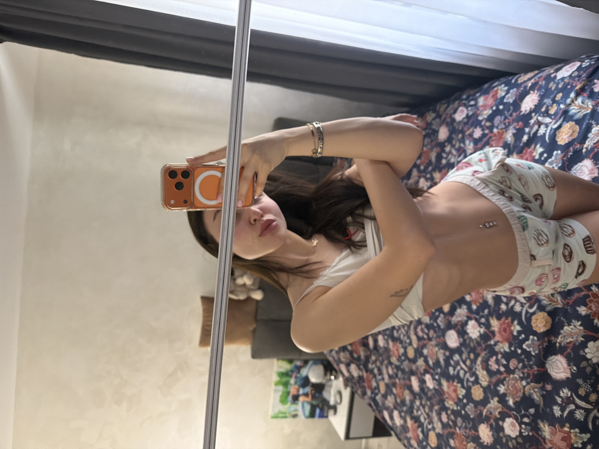 The image displays a mirror selfie taken indoors, specifically within a bedroom environment. A female subject is captured from approximately mid-thigh level upwards, posing in front of a mirror. She has long, dark hair and is attired in a white spaghetti-strap crop top and light-colored shorts adorned with a pattern resembling cupcakes and donuts. Visible personal adornments include a navel piercing (belly button ring), a thin necklace with a small pendant, and two bracelets on her right wrist. A partial tattoo, possibly a stylized wing, is visible on her left upper arm. The subject is holding an orange-cased smartphone, equipped with an orange circular magnetic accessory (likely MagSafe), in her right hand, positioned to capture the reflection.
The background, visible as a reflection in the mirror, reveals a bed with a dark navy blue duvet featuring a floral pattern of pink, orange, and red blooms. Above the bed, there is a dark grey headboard, on top of which rests a brown pillow and a light-colored stuffed animal. To the left of the bed, a nightstand, split between white and a darker color, holds a colorful abstract painting, a dark cylindrical object, and a white, dome-shaped item. The wall behind the bed exhibits a light, textured, off-white or beige finish. To the right of the subject's reflection, a window is partially visible, framed by dark grey and sheer white curtains.
The specific location within the city of Pernik, Bulgaria, is not discernible from the visual information presented in the image.
