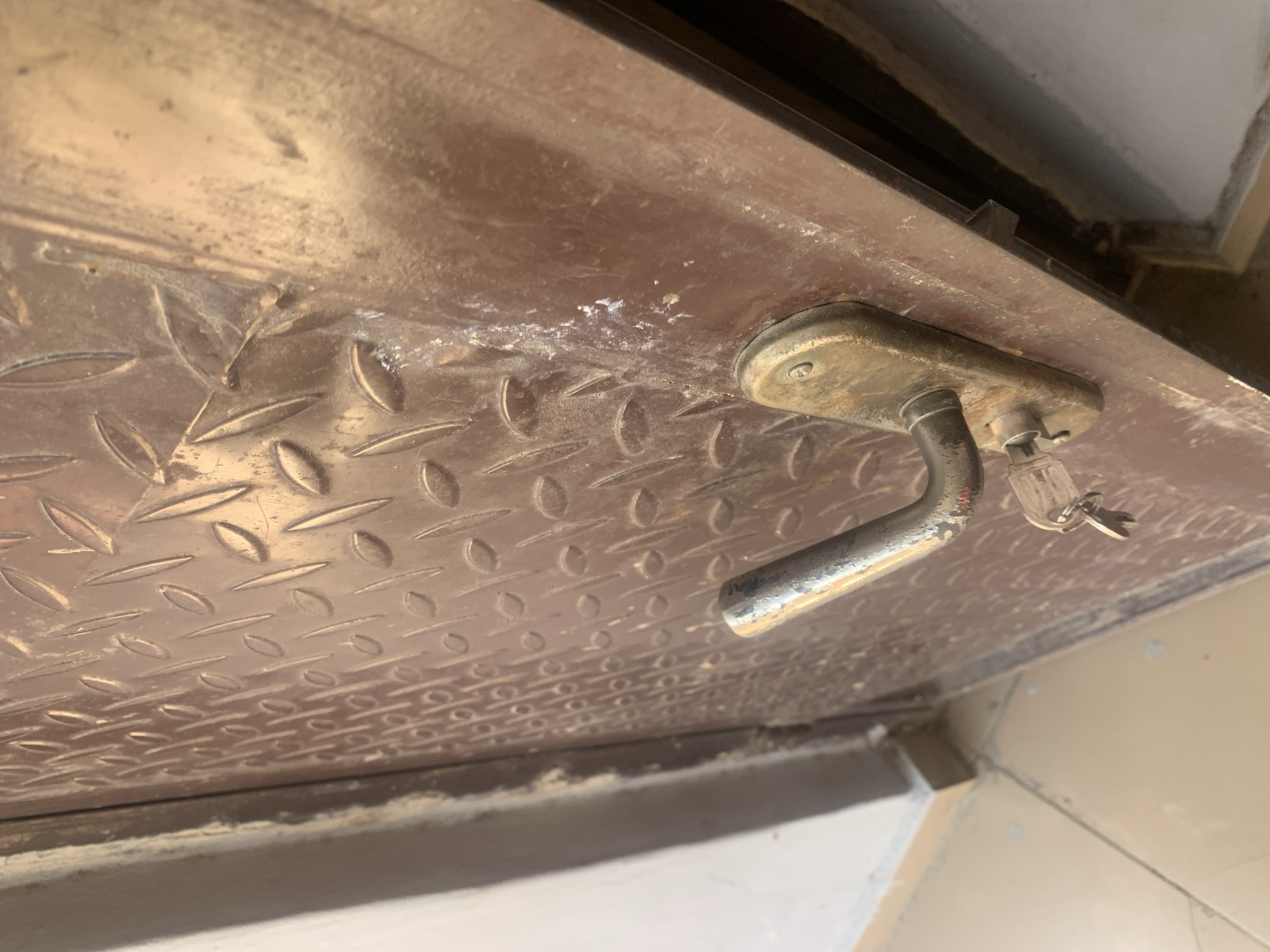 The image shows a close-up view of a brown metal door with a diamond plate texture. A silver door handle with a key in the lock is visible. The door is mounted on a jamb, and the floor in front of it appears to be tiled. The scene is likely an exterior door, possibly in an urban or residential setting in Jauro Yinu, Nigeria, though there are no specific identifiers to confirm this location. The lighting is somewhat harsh, creating reflections on the metallic surfaces. There is a white powdery residue on the door near the handle. The overall impression is of a functional but somewhat aged doorway.