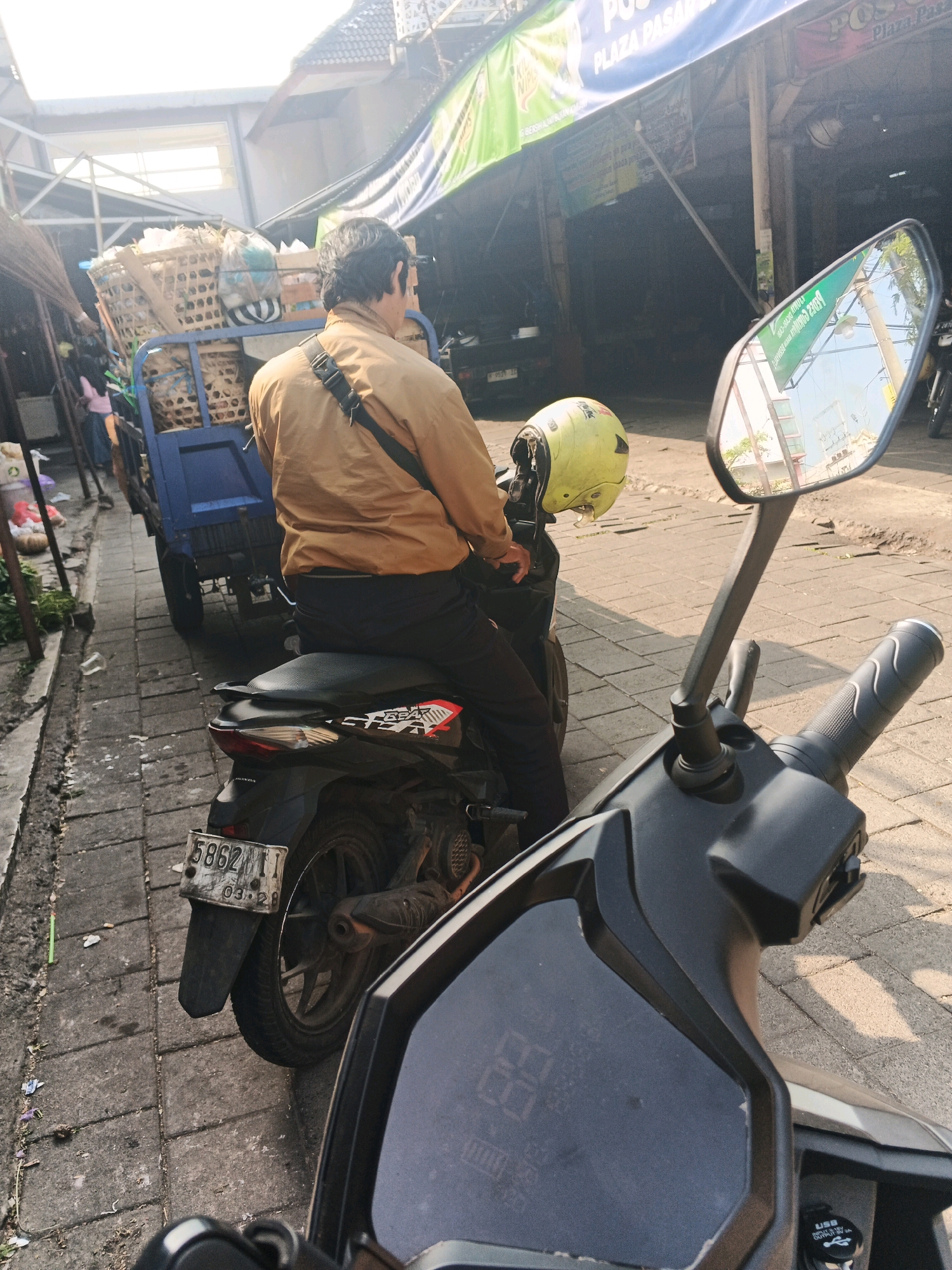 The image depicts a street scene in Ungaran, Indonesia, likely near a market. In the foreground, the handlebars and digital display of a black scooter are visible. A yellow helmet rests on the scooter's seat. A person wearing a tan jacket and dark pants is seated on the scooter, looking towards a blue cart filled with woven baskets. The cart is laden with goods, some of which are wrapped in plastic. The street is paved with gray bricks, and to the left, a sidewalk area is visible with some discarded plastic and debris. In the background, market stalls or buildings with banners are present. The sunlight suggests it is daytime.
